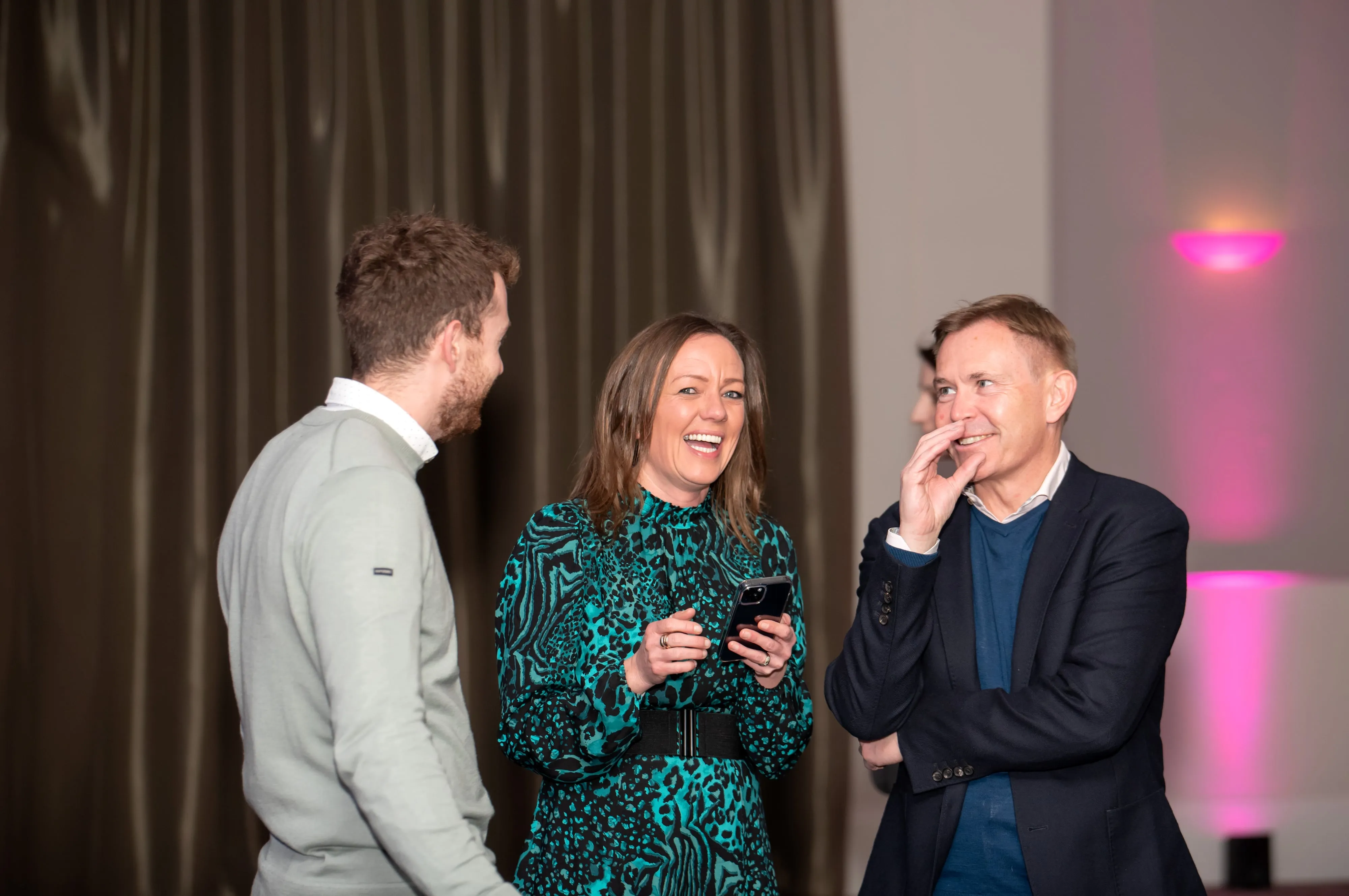 Three colleagues collaborating around a meeting-room table at a corporate workshop, candid laughter mid-conversation