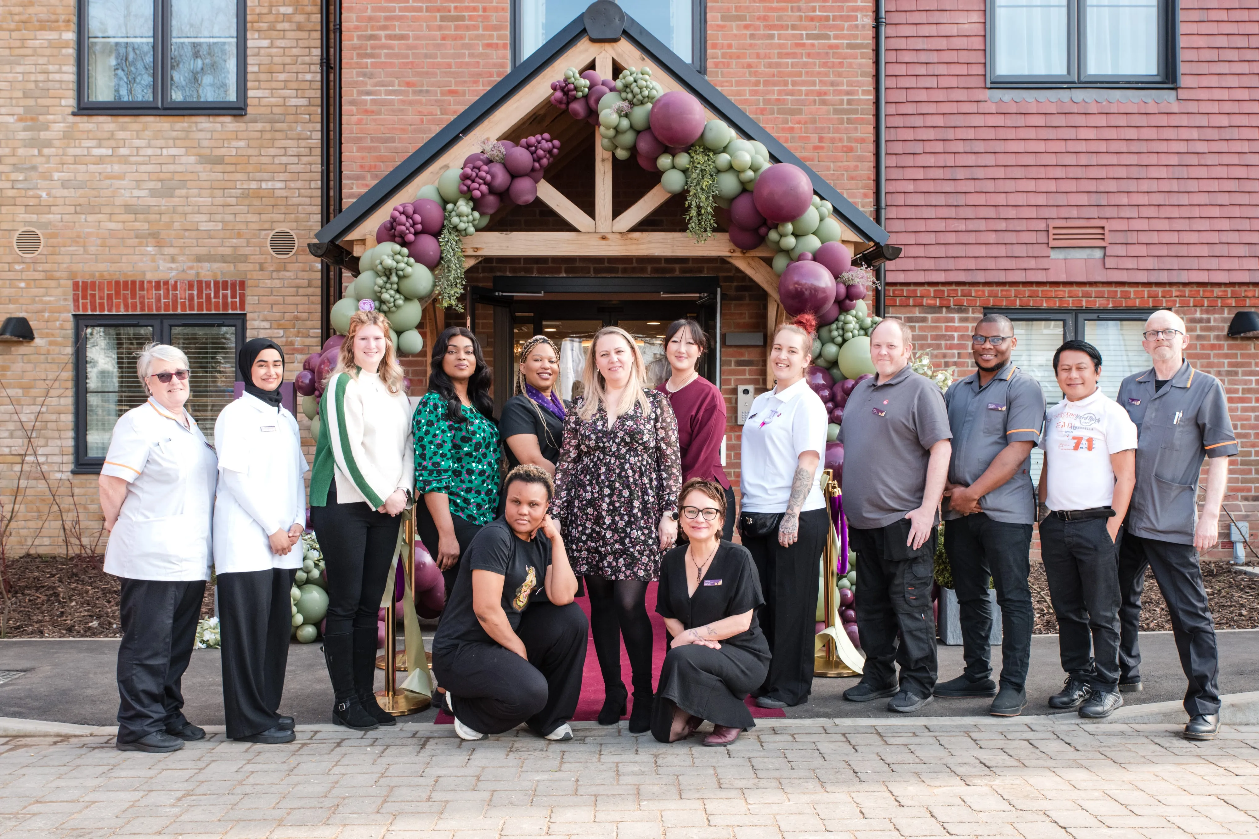 Staff group portrait under a sage-and-lilac balloon arch at a care-home opening event