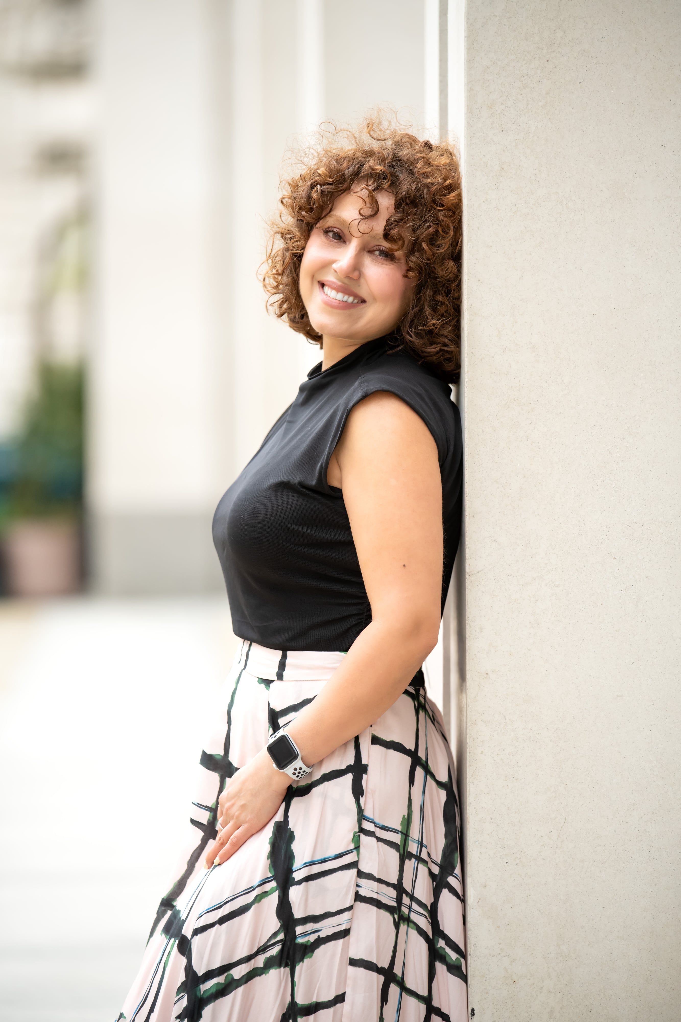 Personal-brand portrait — woman in black top and plaid maxi skirt leans against a stone pillar on a London street