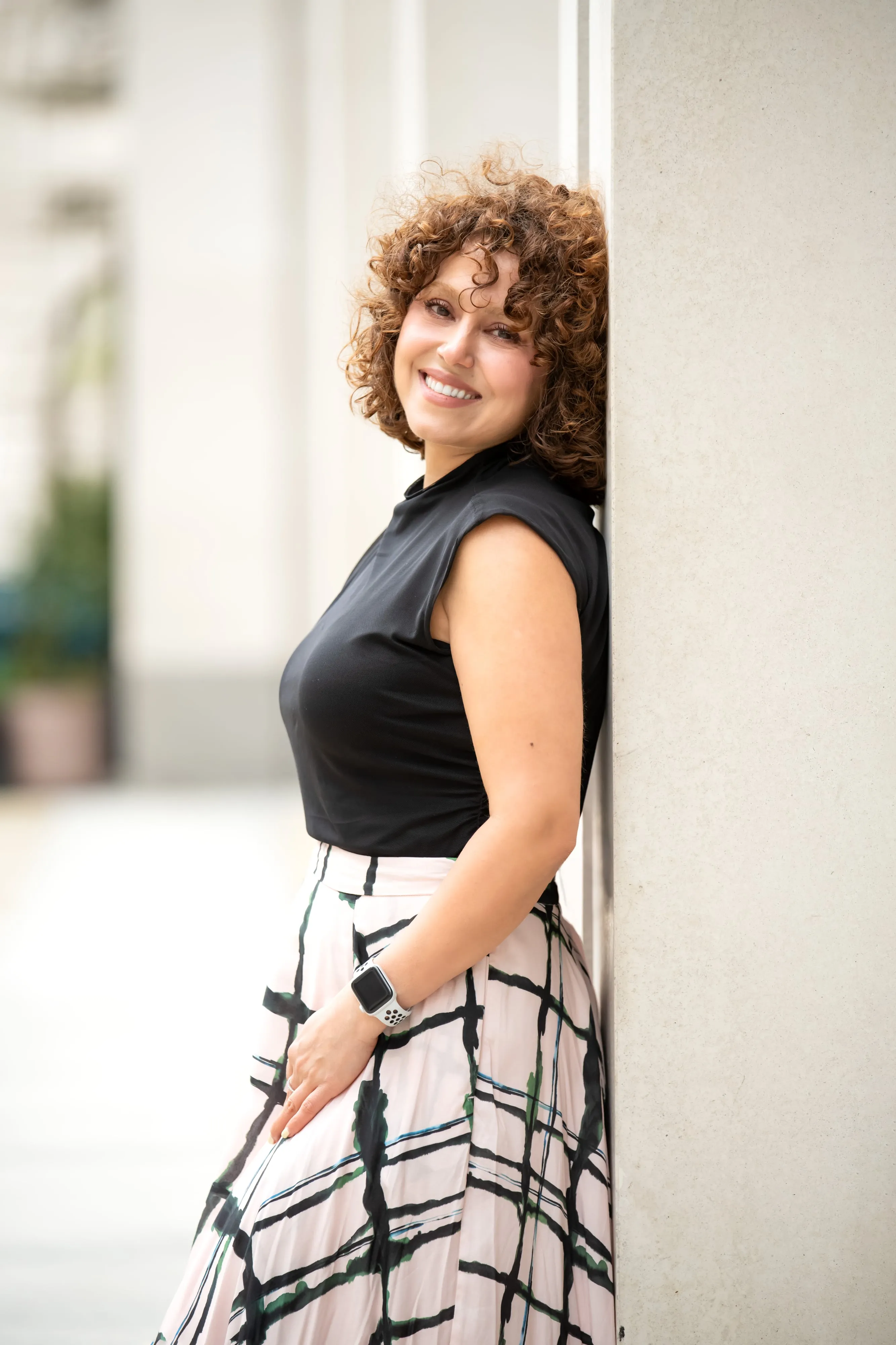 Personal-brand portrait — woman in black top and plaid maxi skirt leans against a stone pillar on a London street