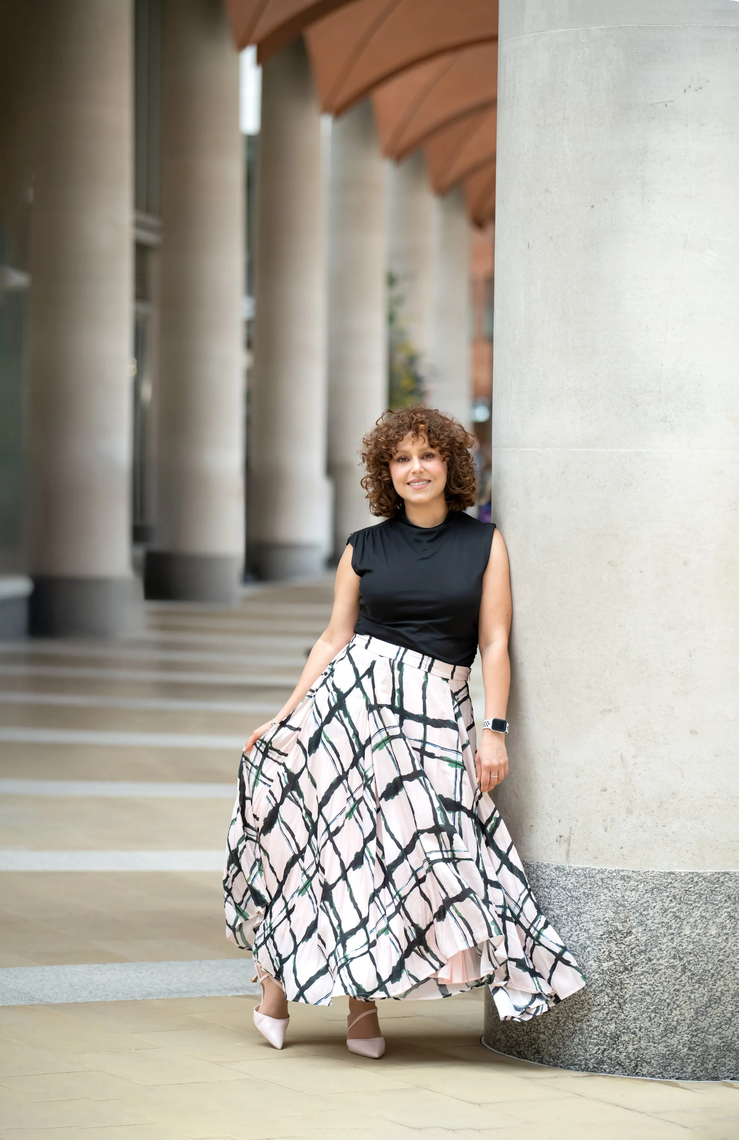 Personal-brand portrait under a London colonnade — woman in black top, plaid maxi skirt held wide, arched ceiling overhead