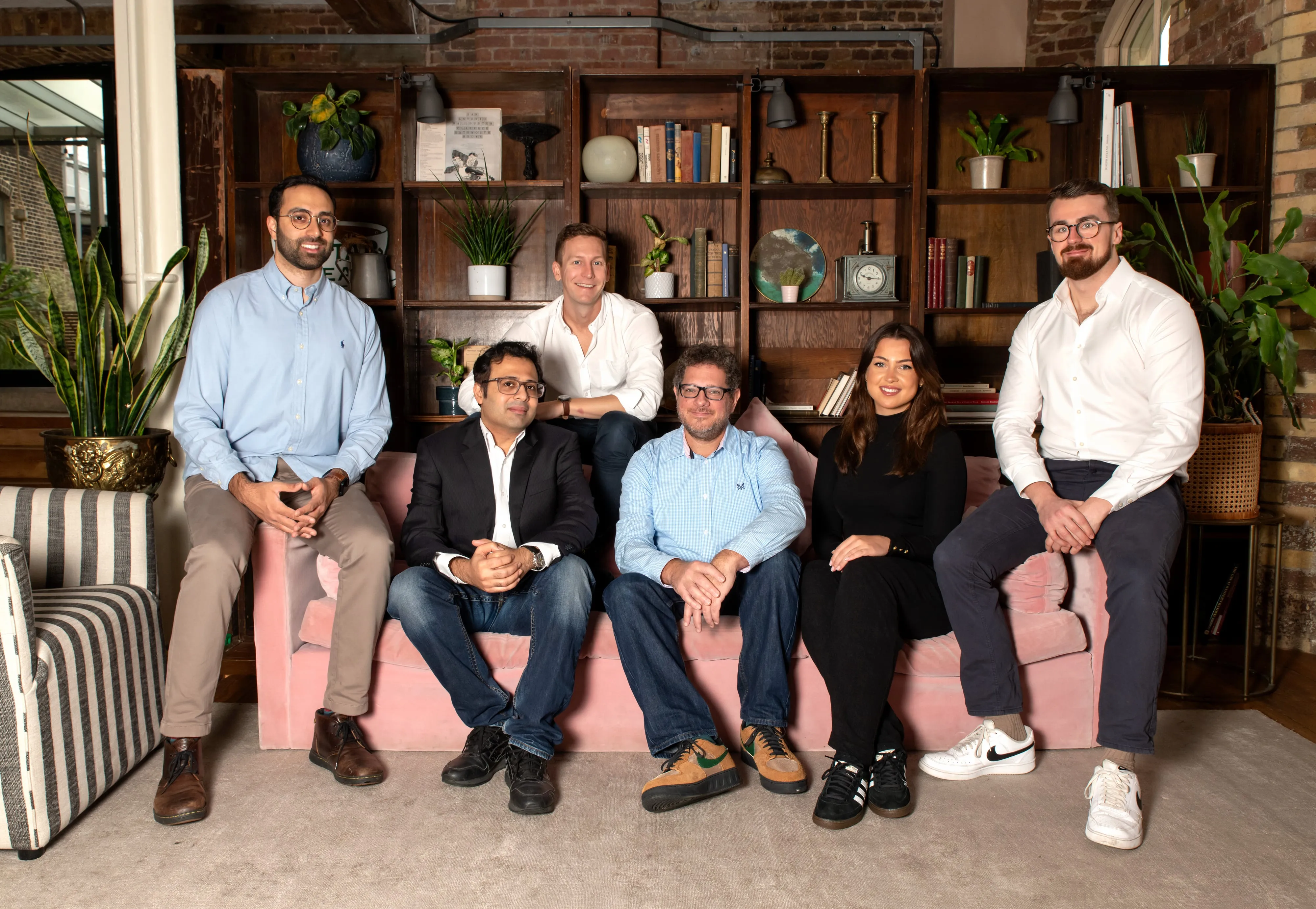 Sprout AI team member solo portrait on a stool by the loft windows, white shirt and glasses, brick wall and pendant lights behind