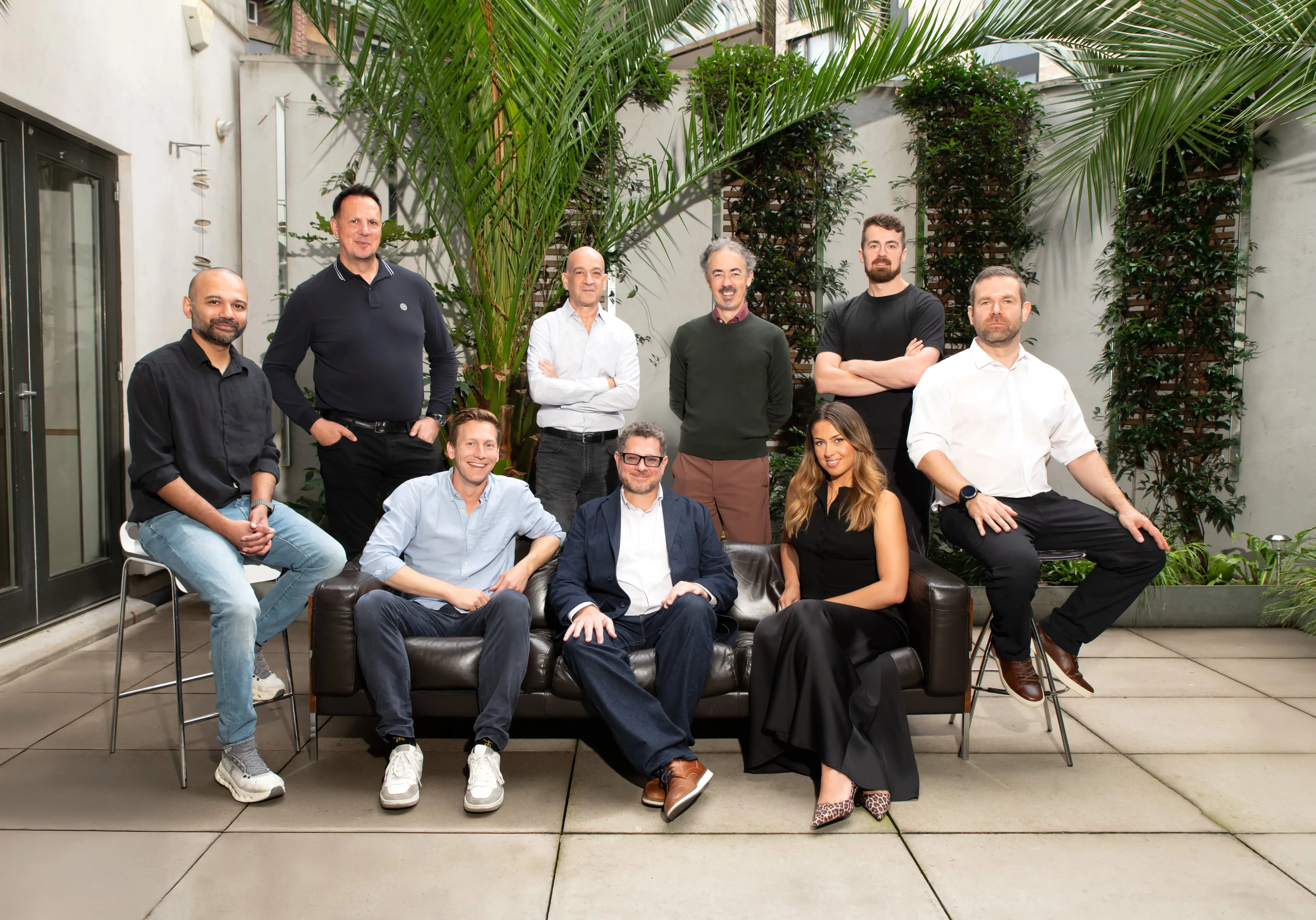 Sprout AI team of nine in a courtyard portrait around a black leather sofa, palms and climbing greenery behind