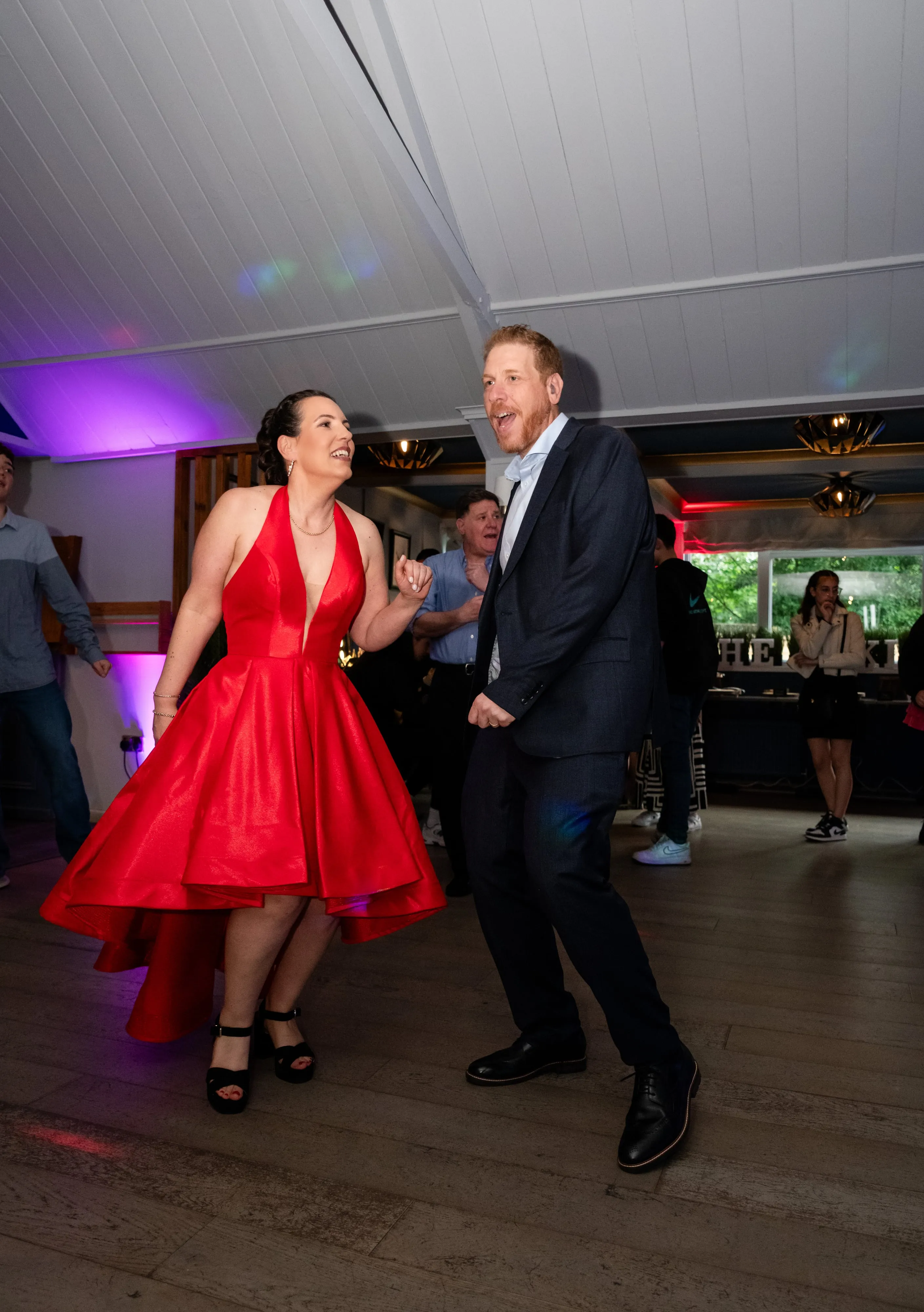 Mum in red gown laughs as she dances with a guest in suit on the dance floor under stage lights