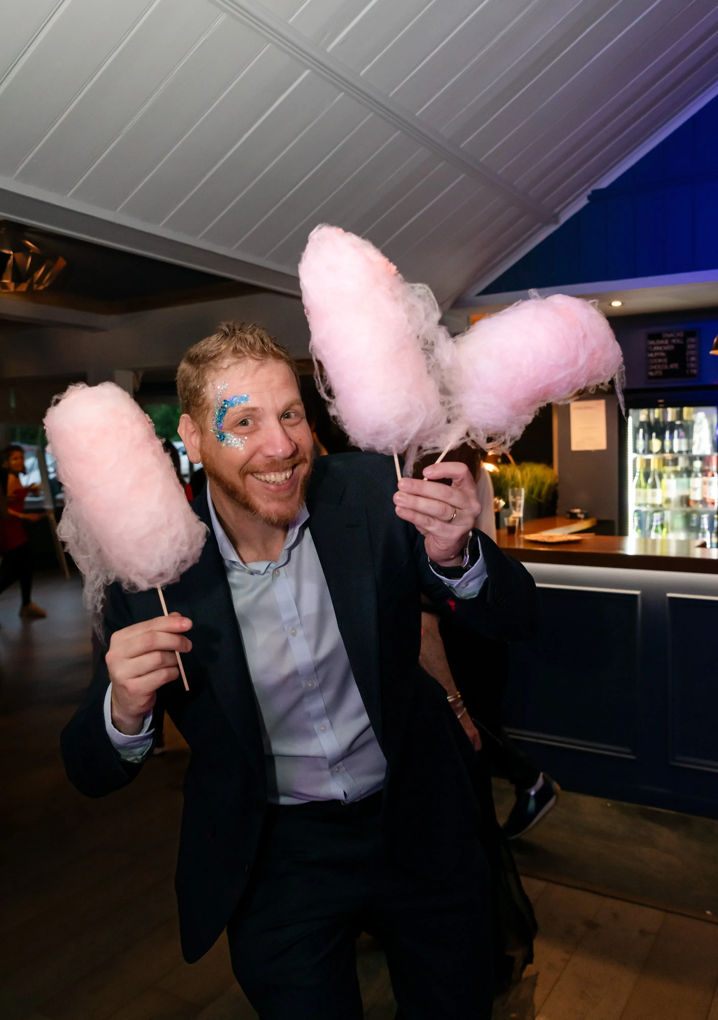 Adult guest in dark suit with sequinned face decoration grins holding two pink candy-floss sticks at the venue bar
