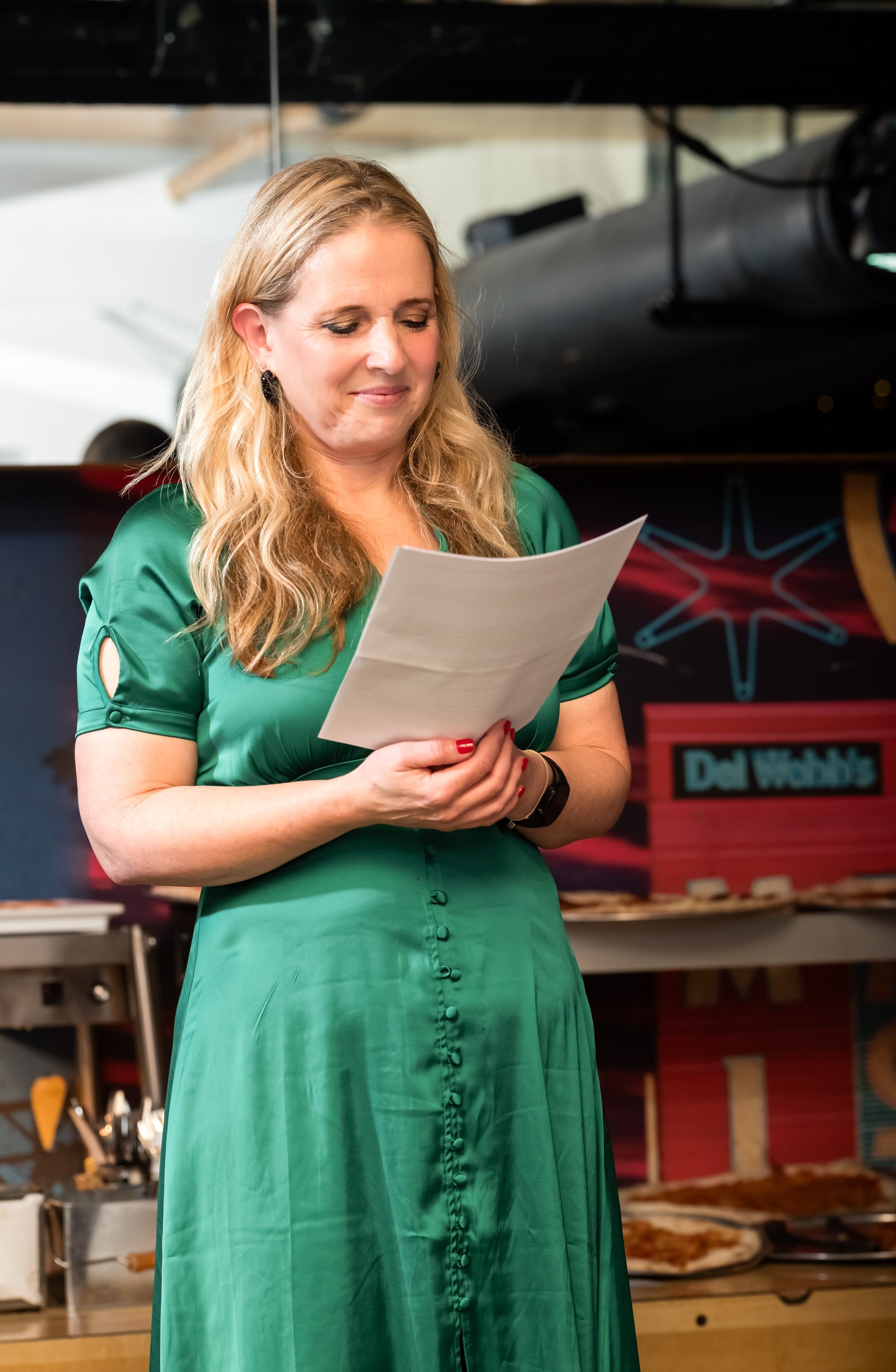 Mum in green satin dress reads a heartfelt speech into a microphone at the party venue stage