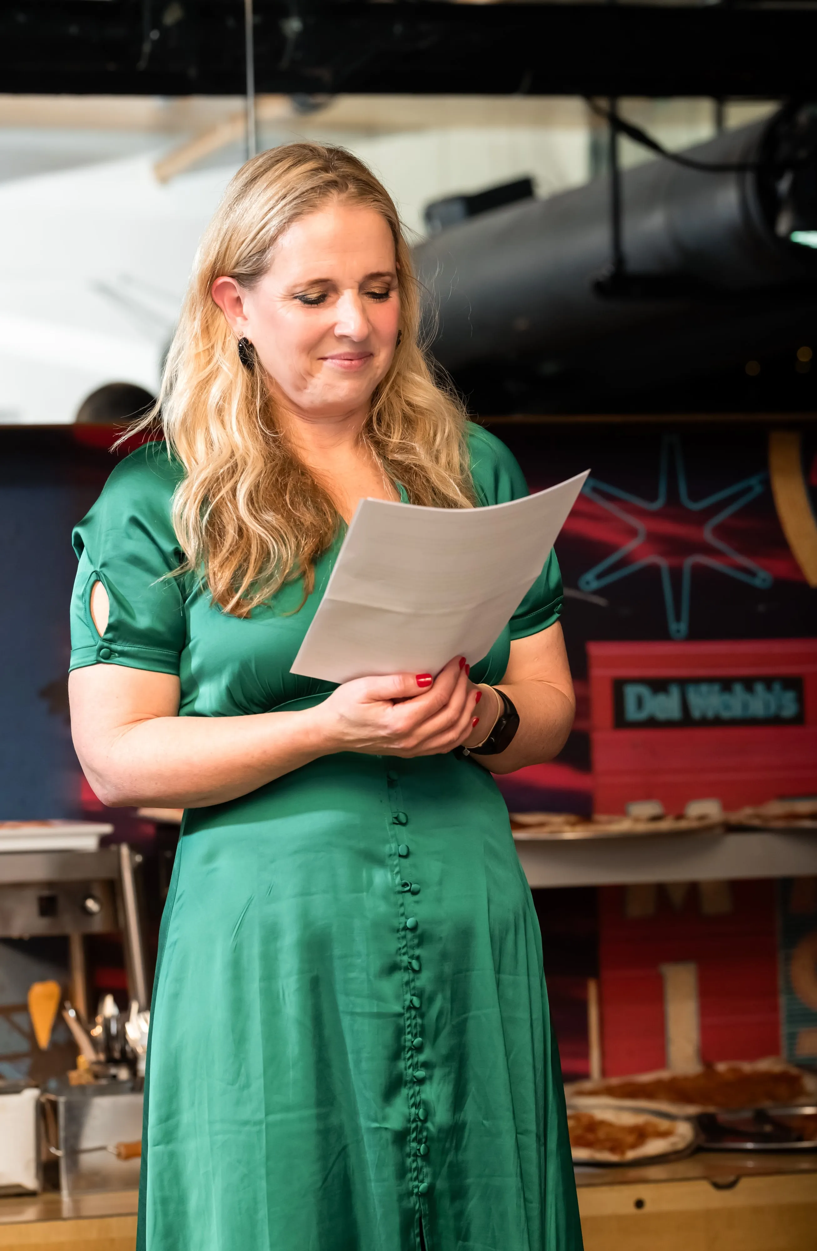 Mum in green satin dress reads a heartfelt speech into a microphone at the party venue stage