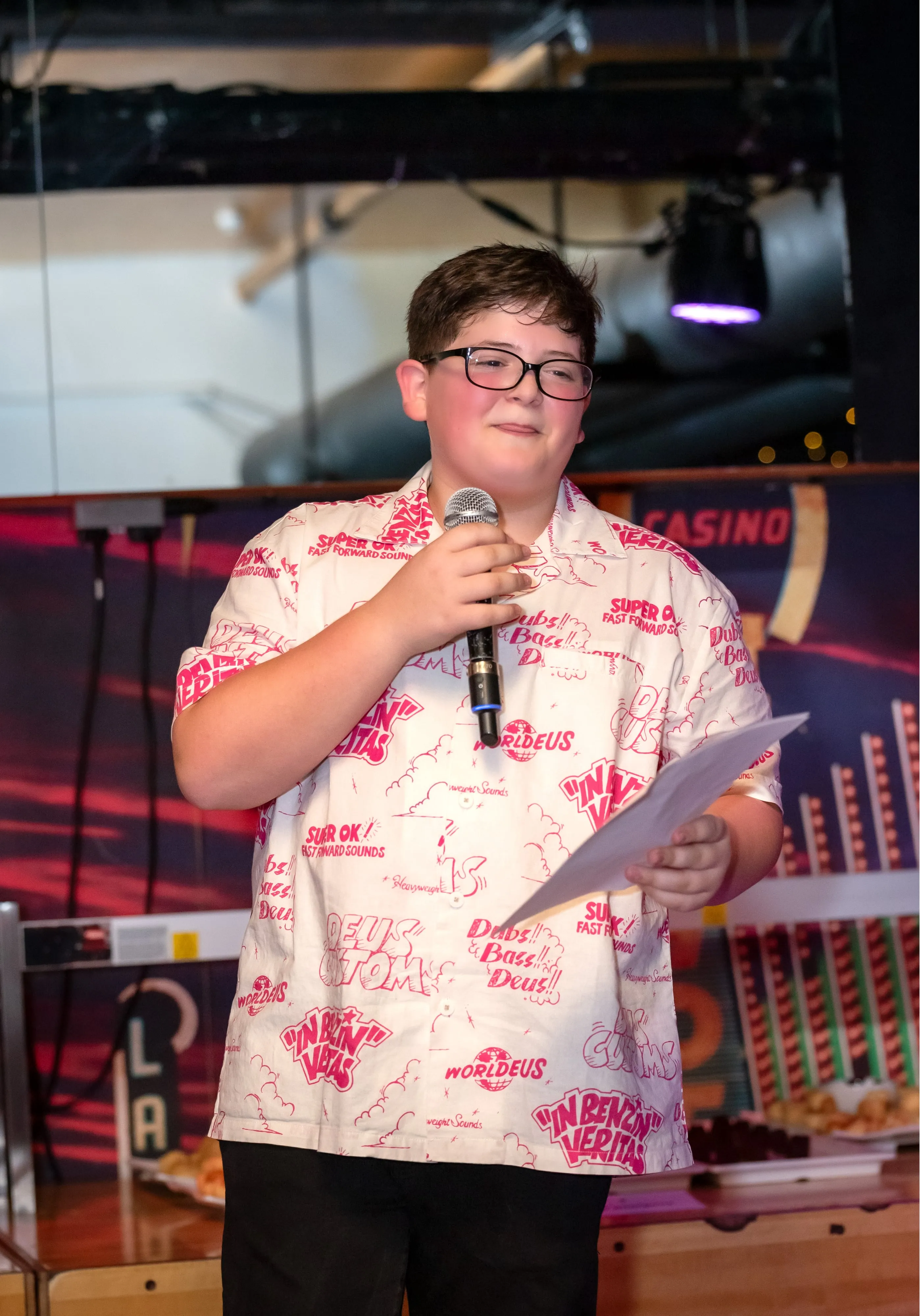 Bar mitzvah boy in printed shirt and glasses gives his speech holding a microphone and notes on the stage