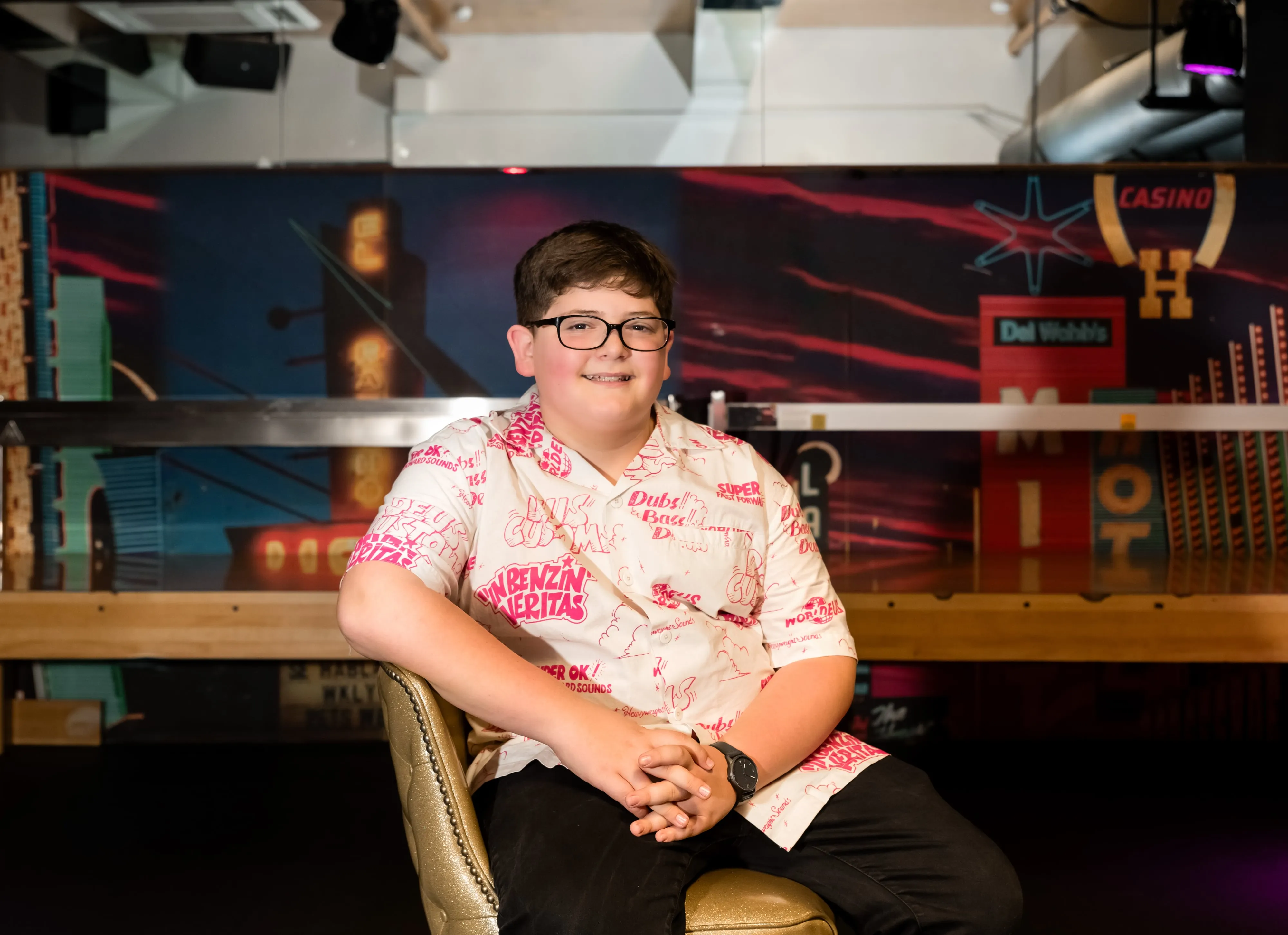 Bar mitzvah boy in printed shirt and glasses sits relaxed in a gold armchair against a neon-painted Vegas backdrop