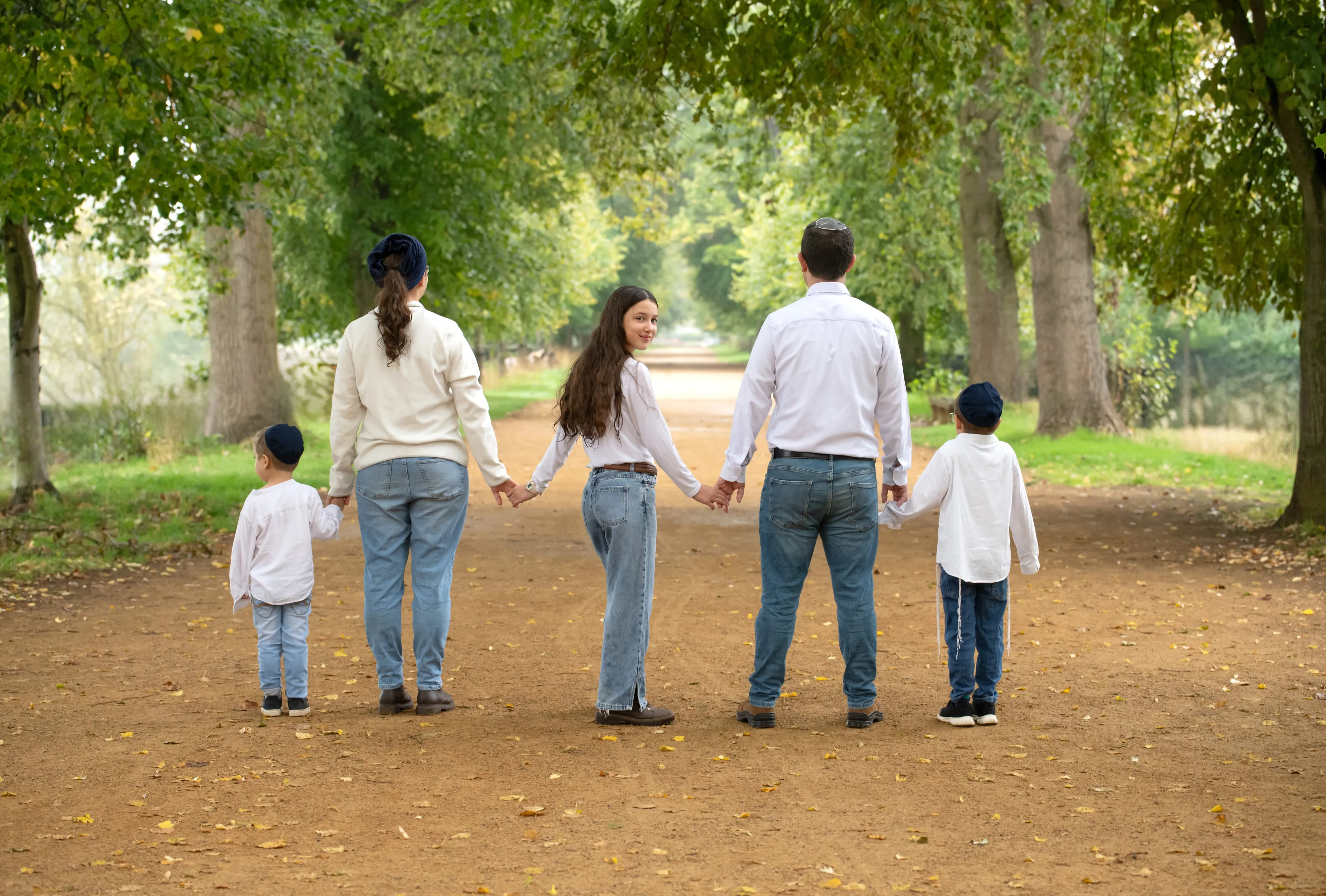 Family of five hold hands on a tree-lined autumn path, all in white shirts