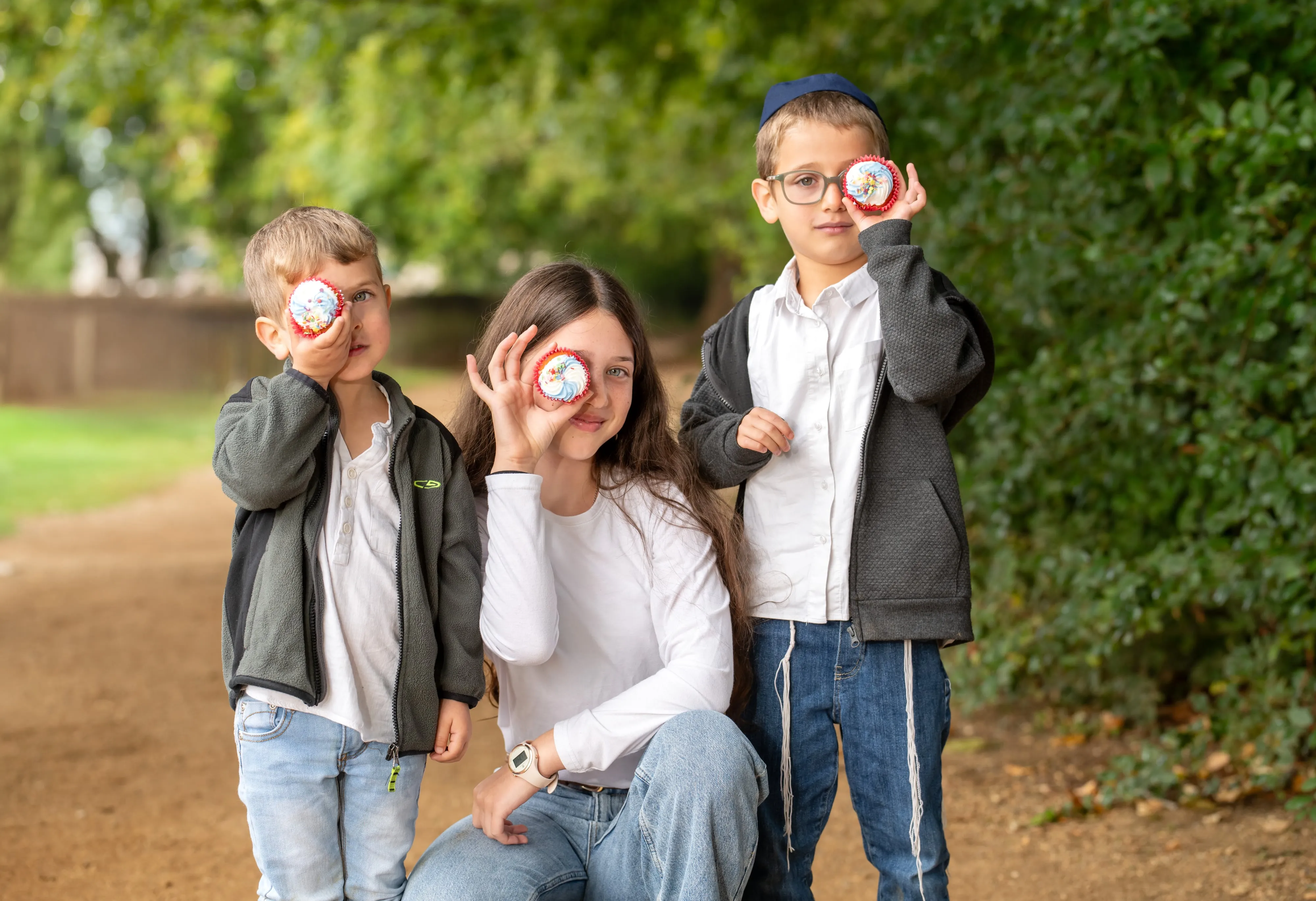 Bat mitzvah girl and her two younger brothers in kippot peek through brightly iced cupcakes held up as eye-pieces in dappled parkland