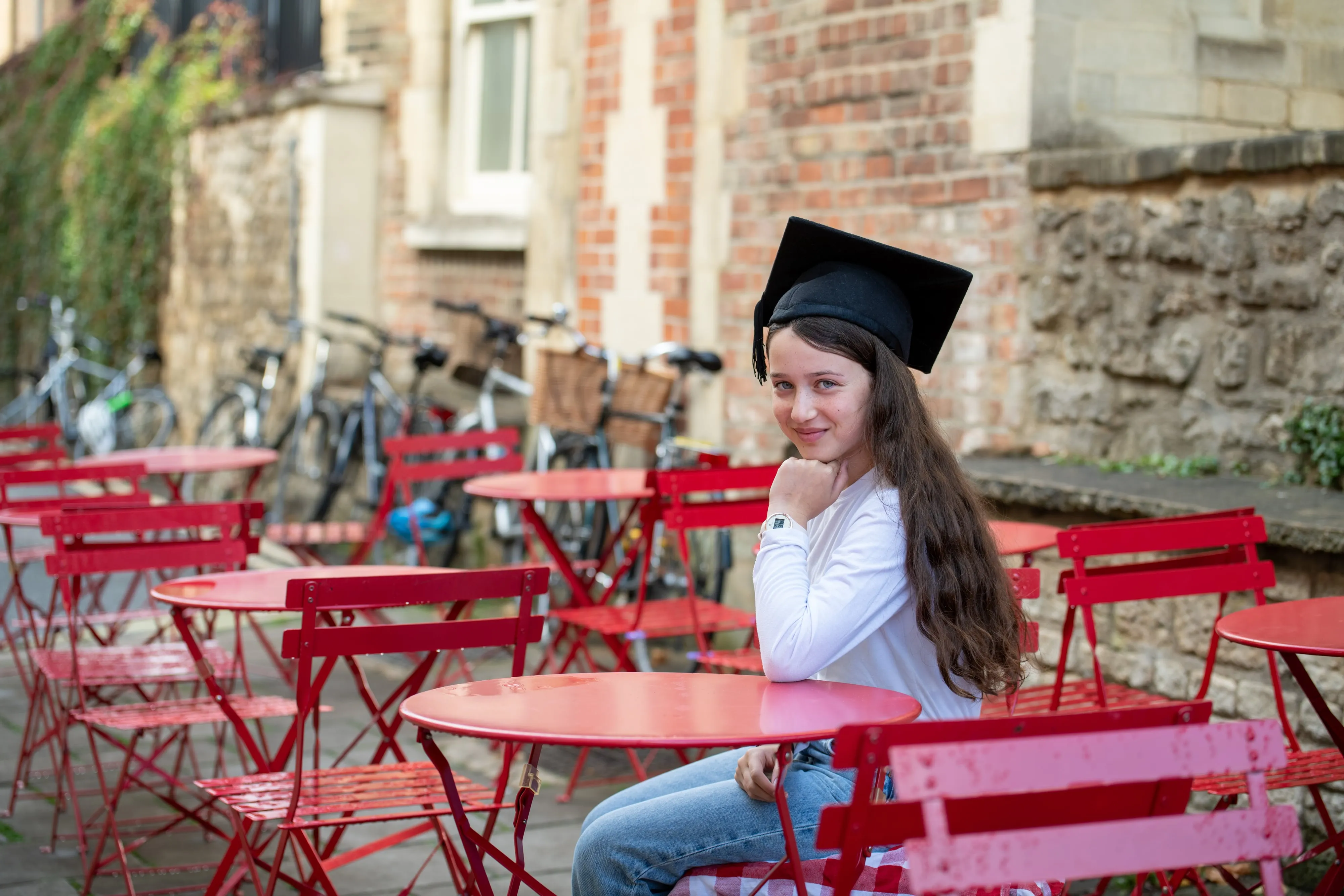 Bat mitzvah girl wears a graduation mortar board at a sunny Oxford courtyard with red bistro chairs