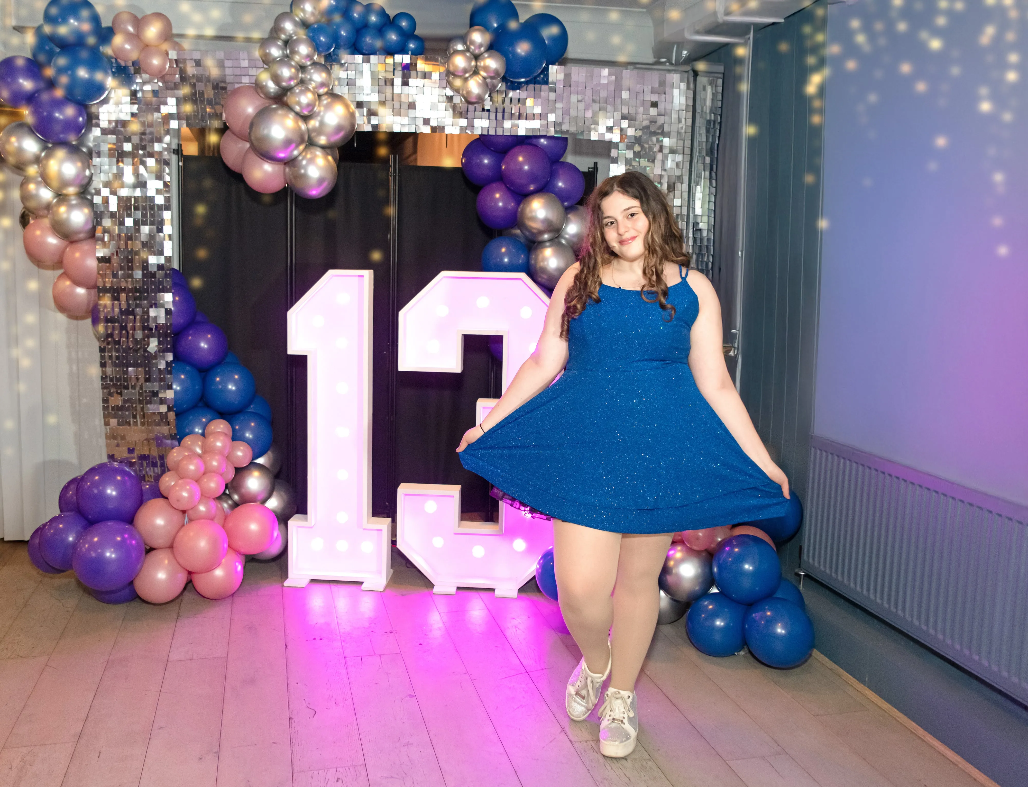 Bat mitzvah girl twirls her sparkly blue dress beside a giant illuminated 13 and balloon disco arch