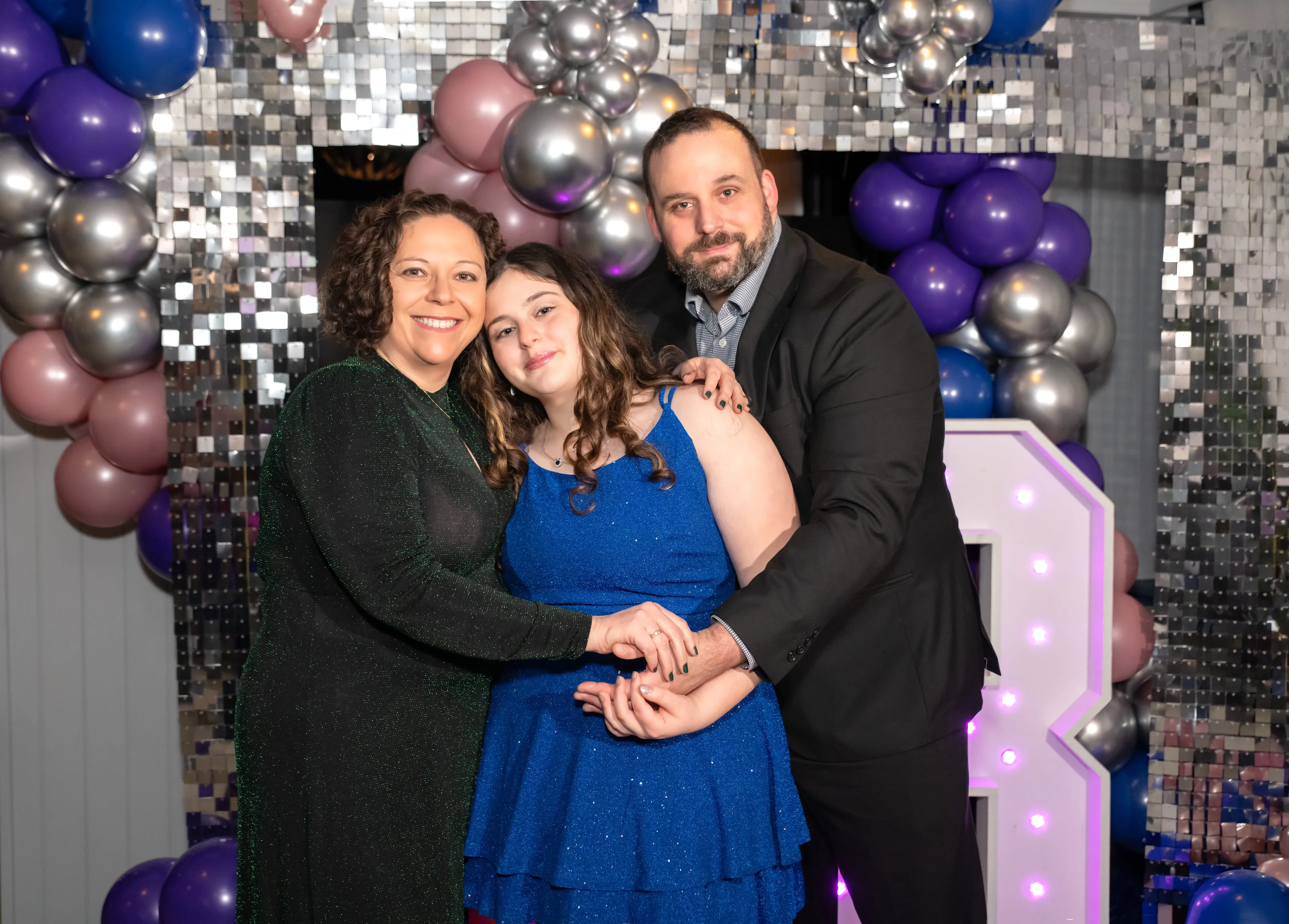Mum in green satin, bat mitzvah daughter in royal-blue sequins and dad in dark suit share a warm embrace under a balloon and silver-sequin disco arch