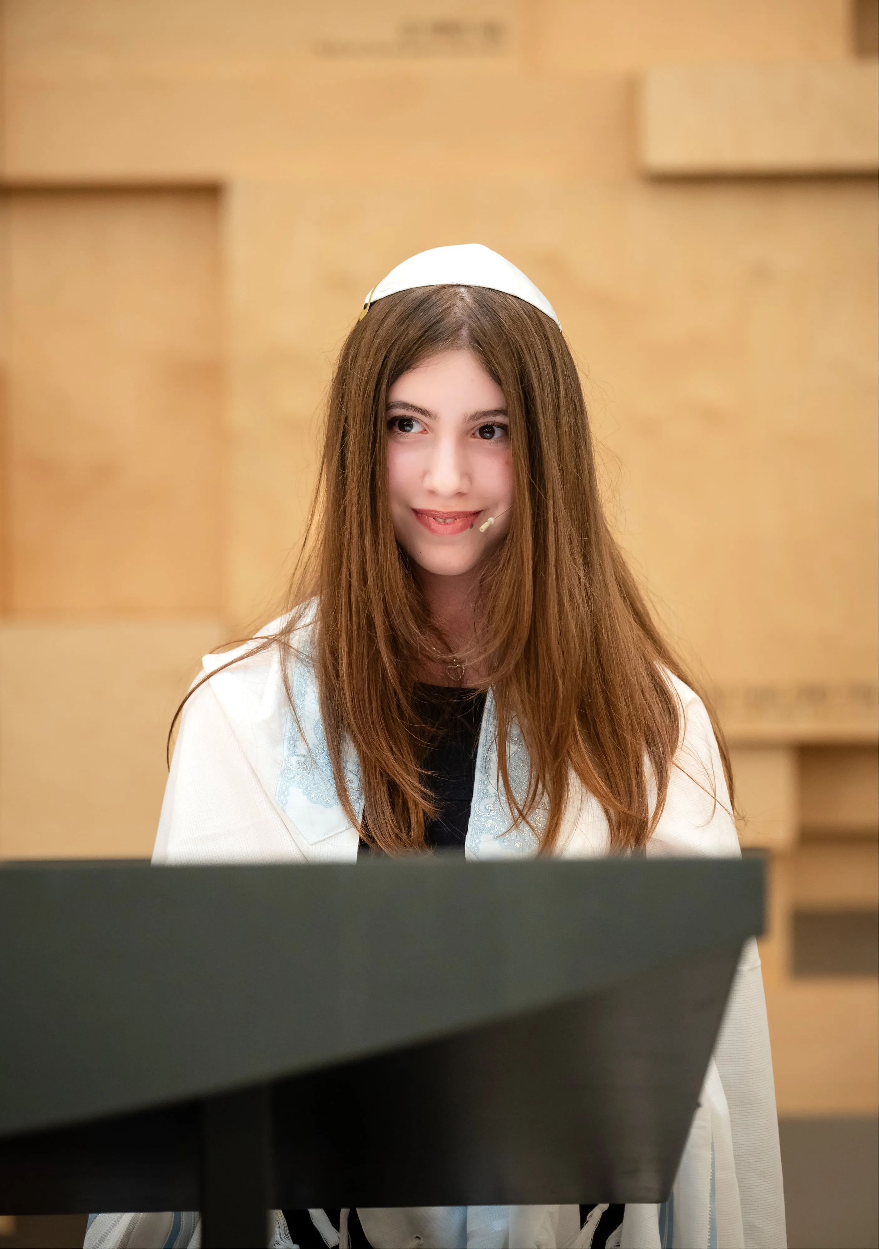 Bat mitzvah girl in white kippah and tallit smiles at the bimah against pale-wood synagogue panelling
