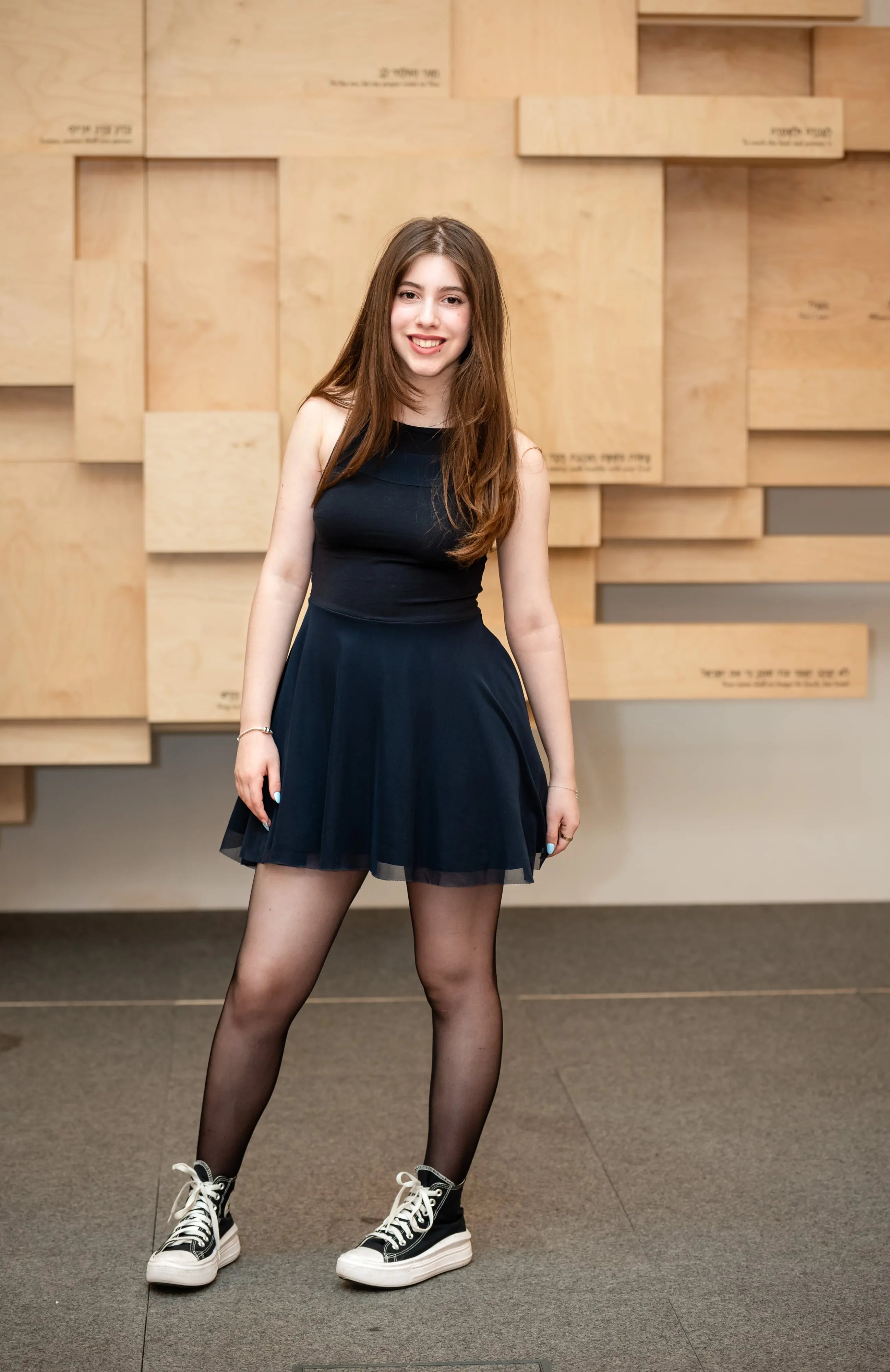 Bat mitzvah girl in navy dress and trainers smiles against the Hebrew-inscribed wooden block wall of the synagogue