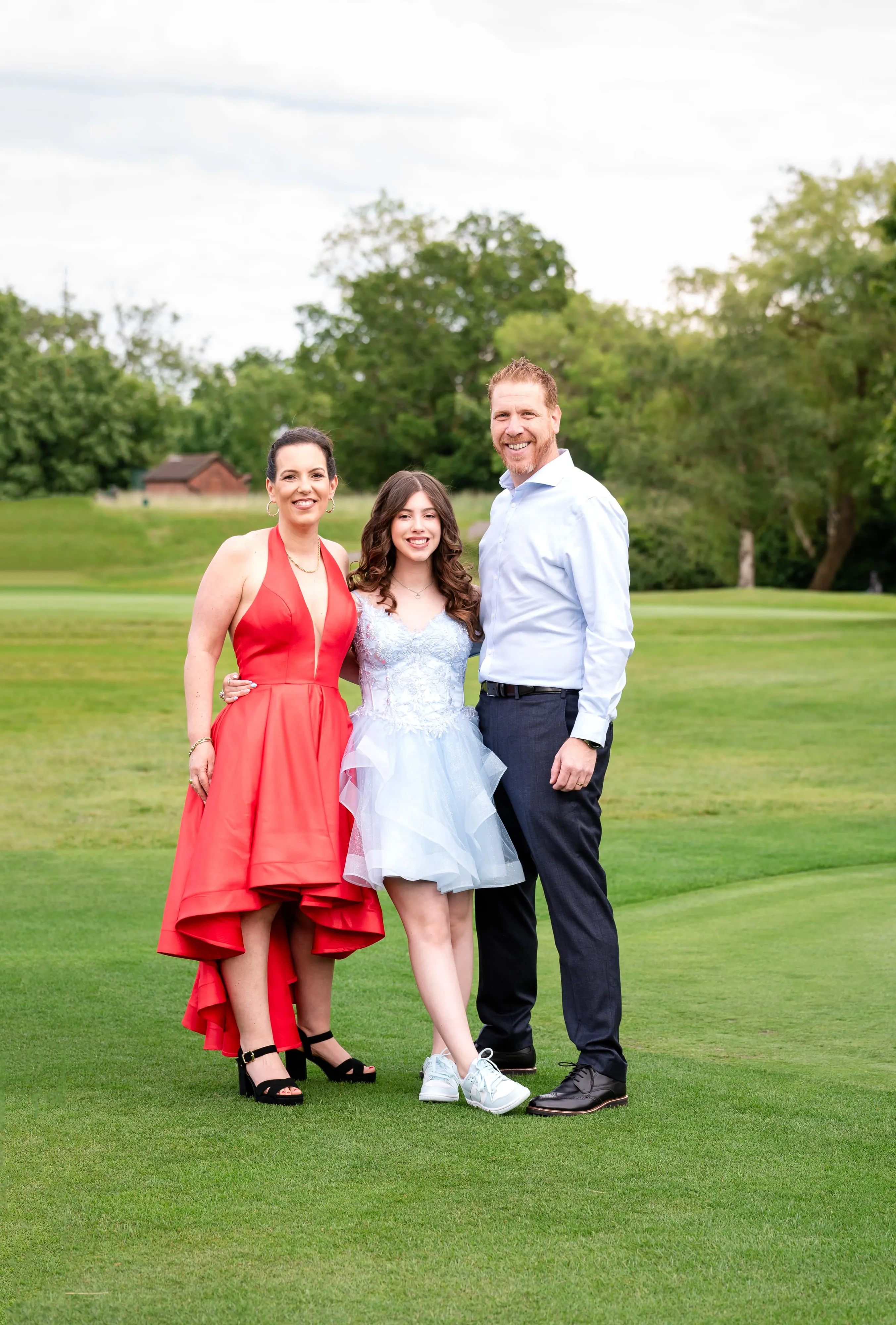 Bat mitzvah family of three pose together on the golf-course lawn, mum in red, daughter in pale blue