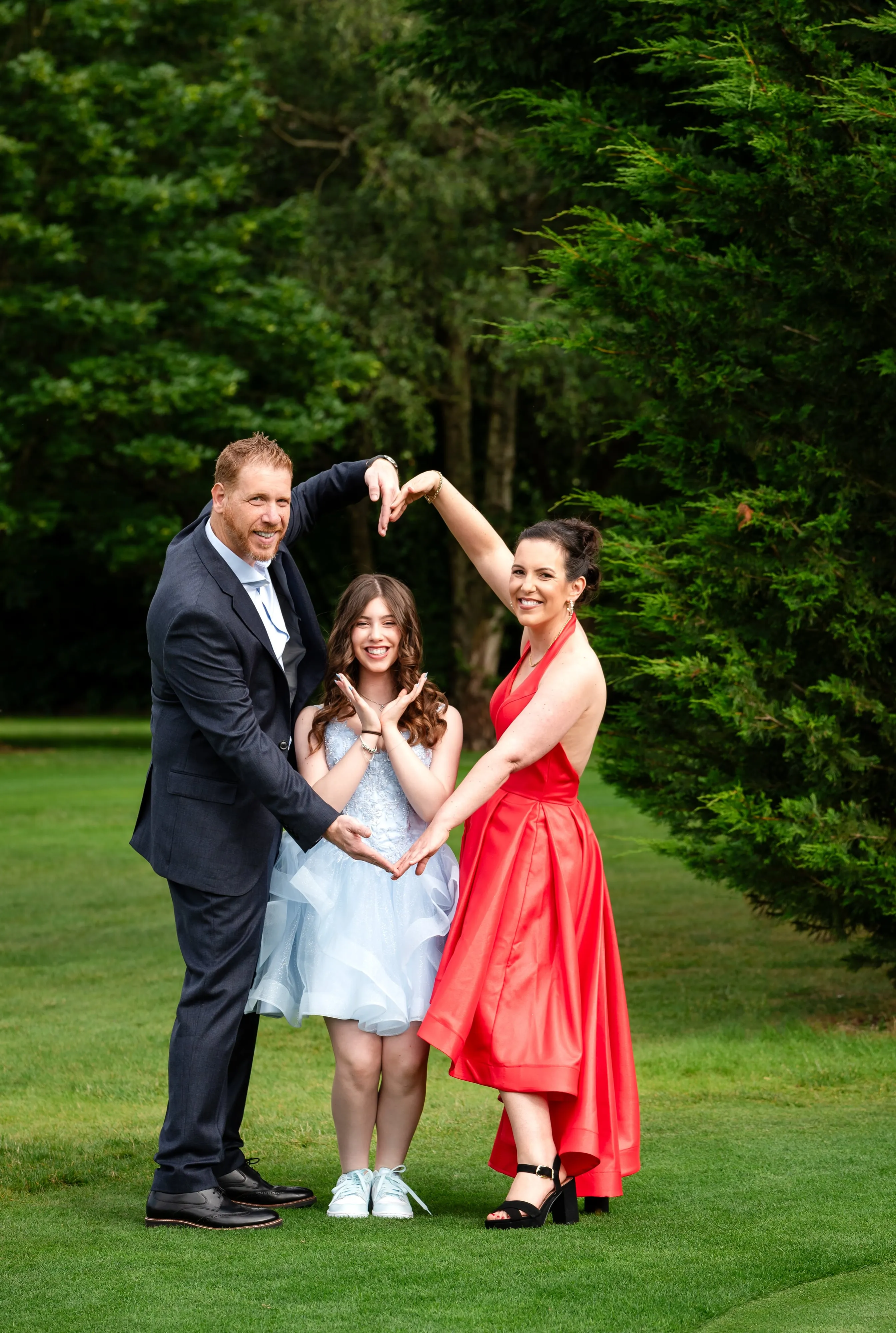 Bat mitzvah parents form a heart shape with their arms above their daughter in pale-blue dress on the golf-course lawn