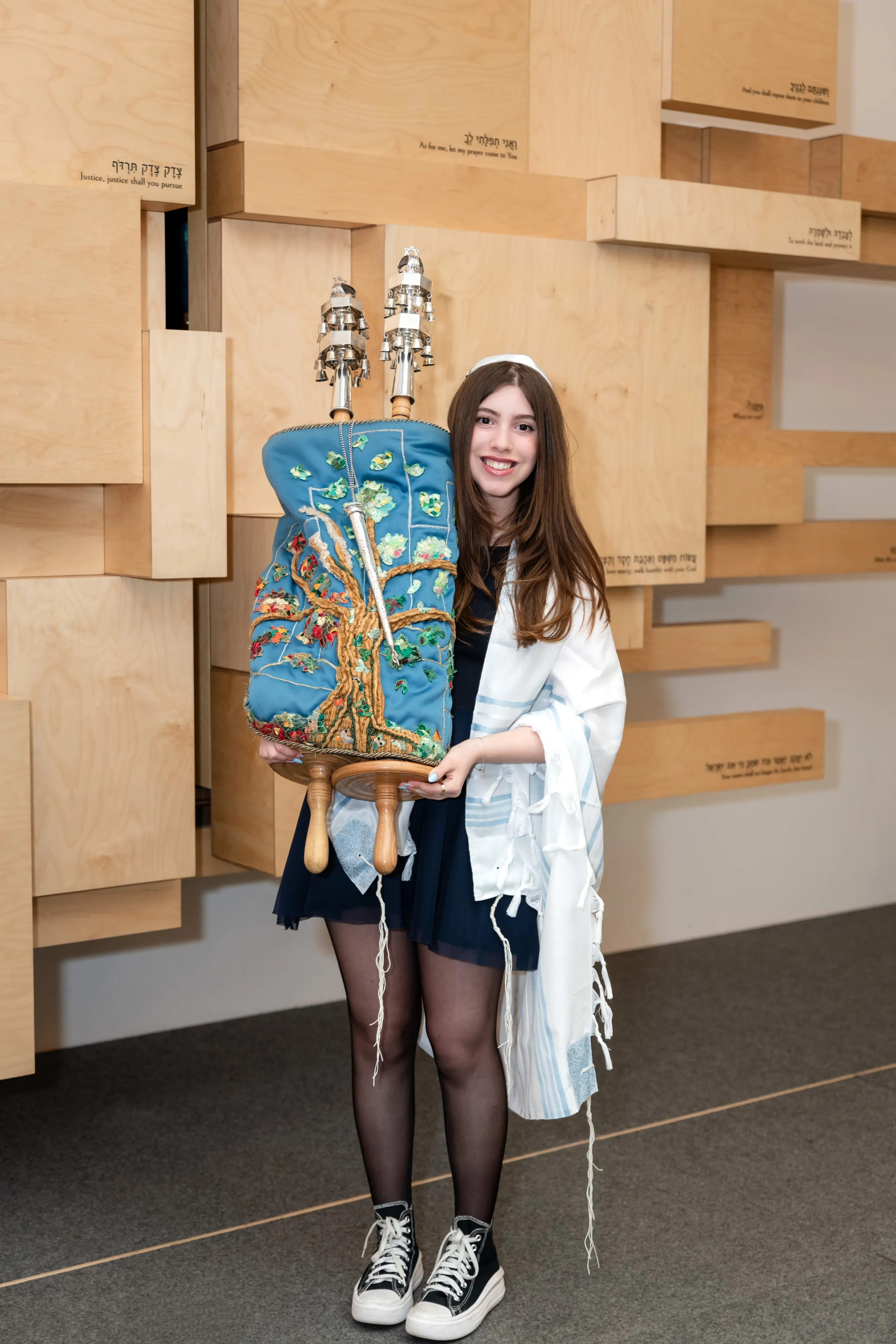 Bat mitzvah girl in tallit holds the embroidered Torah scroll proudly against the synagogue plaque wall