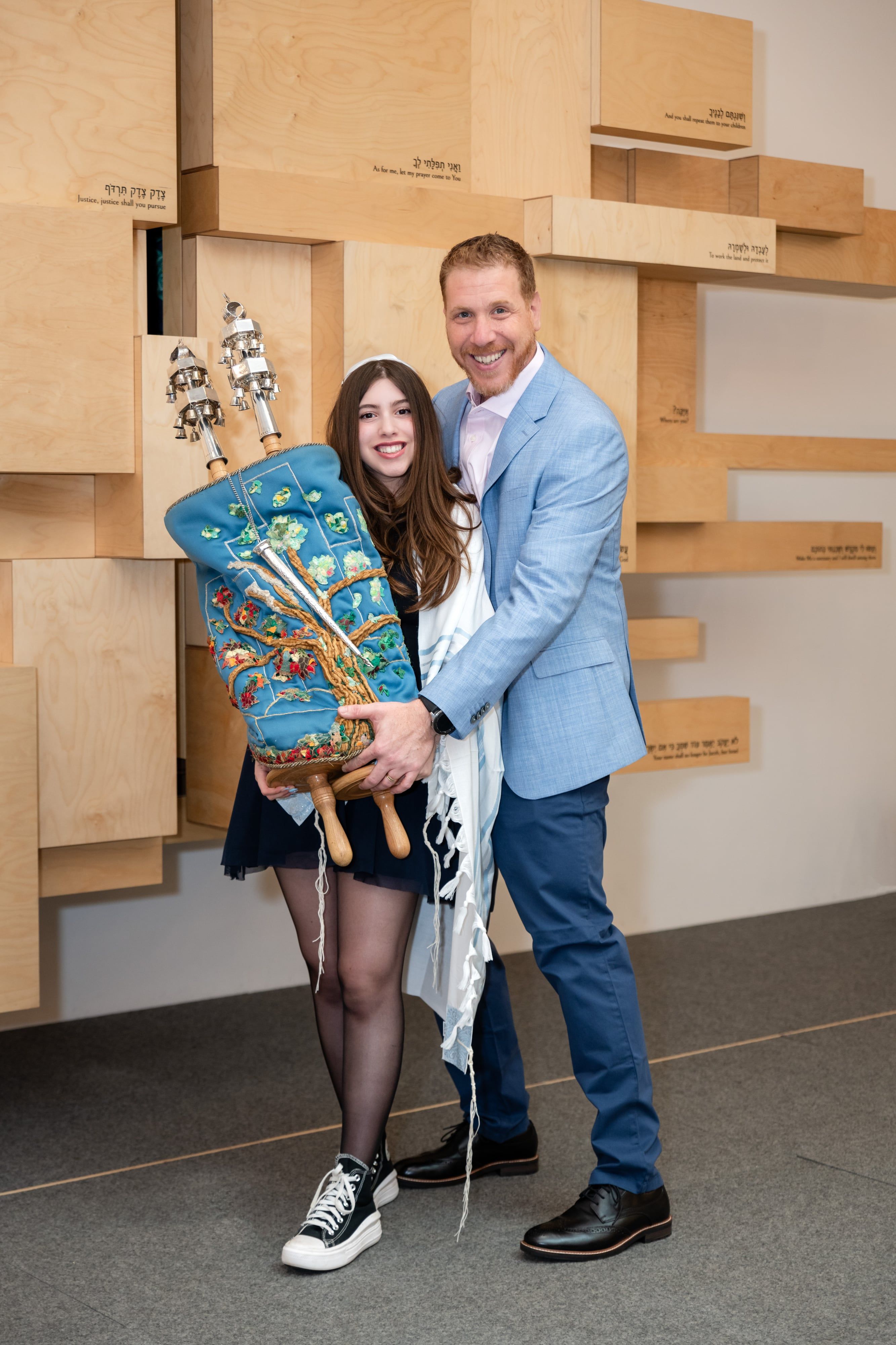 Bat mitzvah girl and her father in pale-blue suit hold the embroidered Torah scroll together against the Hebrew-inscribed synagogue wall