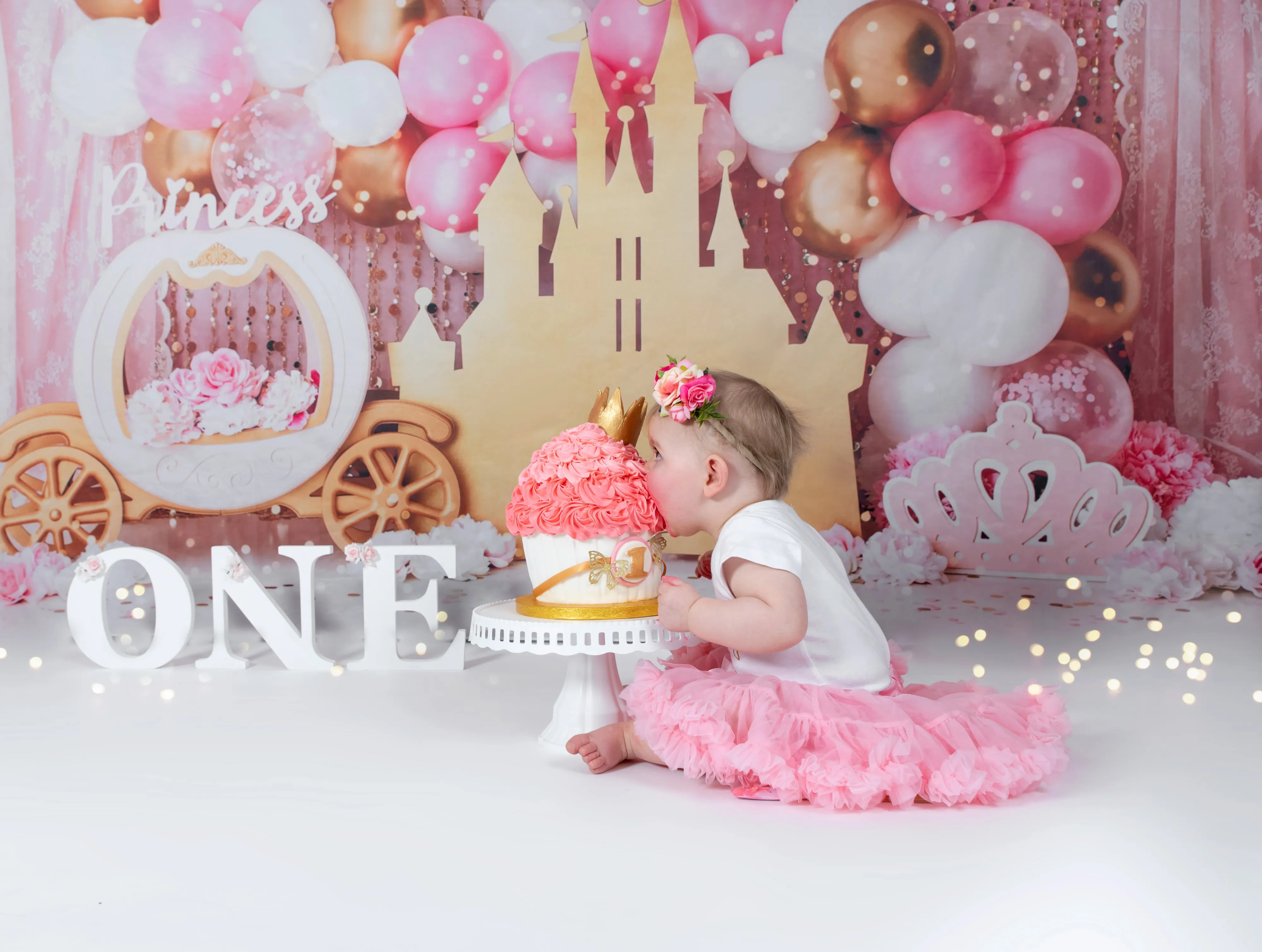 Toddler in pink tutu kisses pink ruffle cake on princess castle set with carriage and balloons