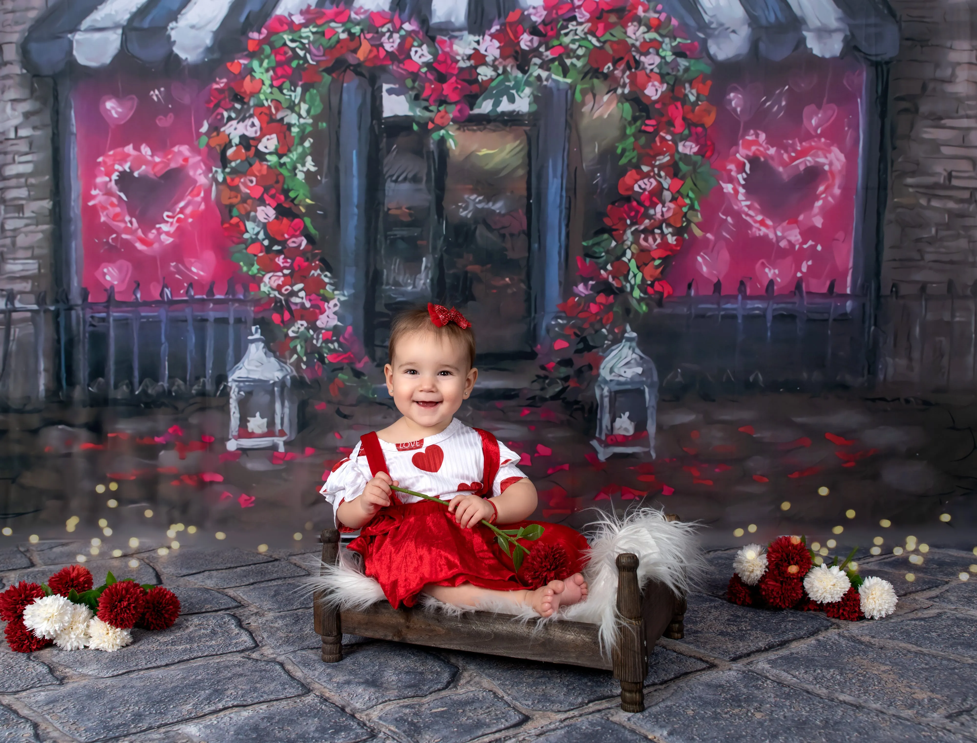 Toddler in red tutu sits on wooden bed in Valentines set with red rose archway and pom-poms