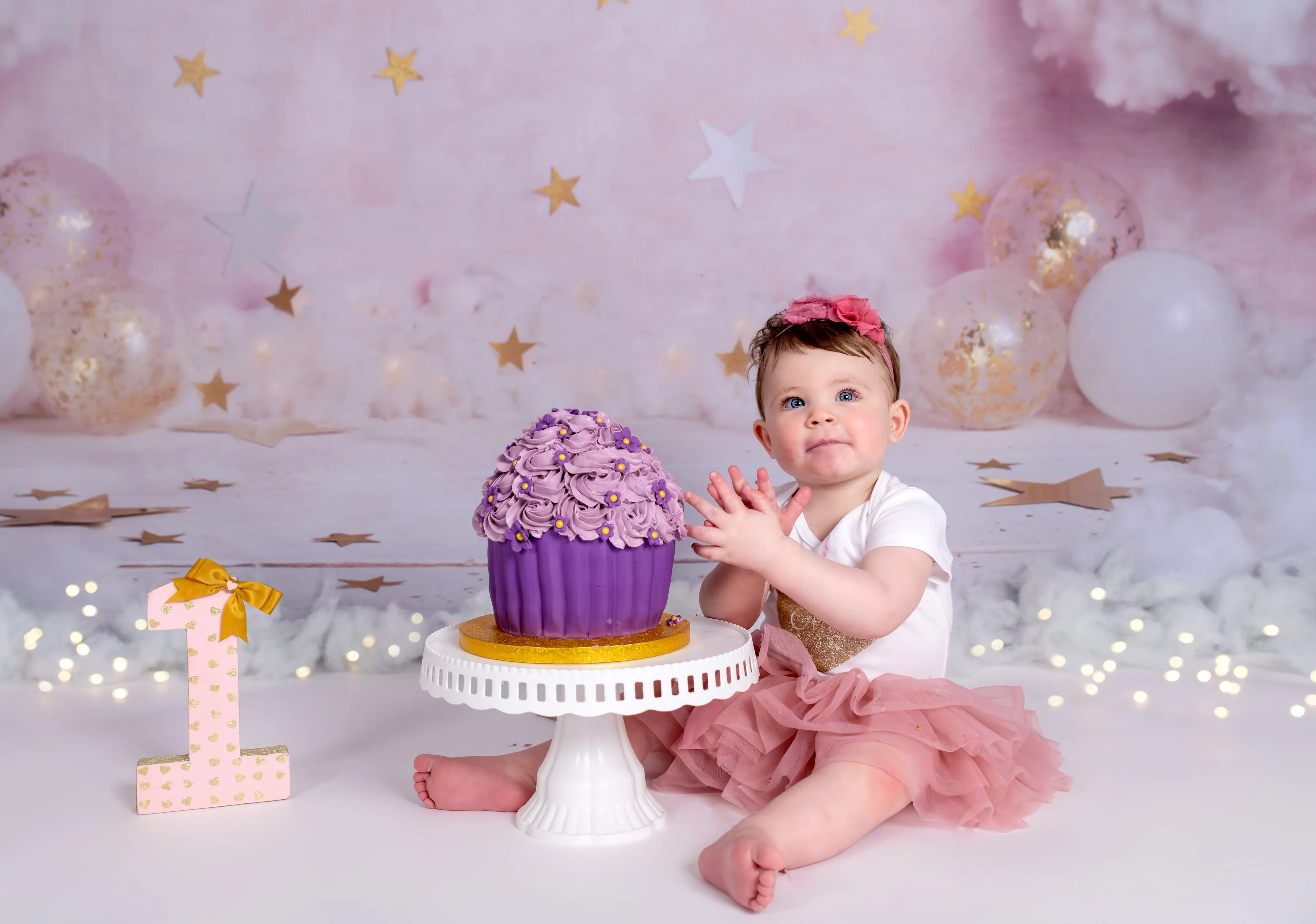 Toddler in pink tutu touches purple giant cupcake with gold 1 on pink stars-and-clouds set
