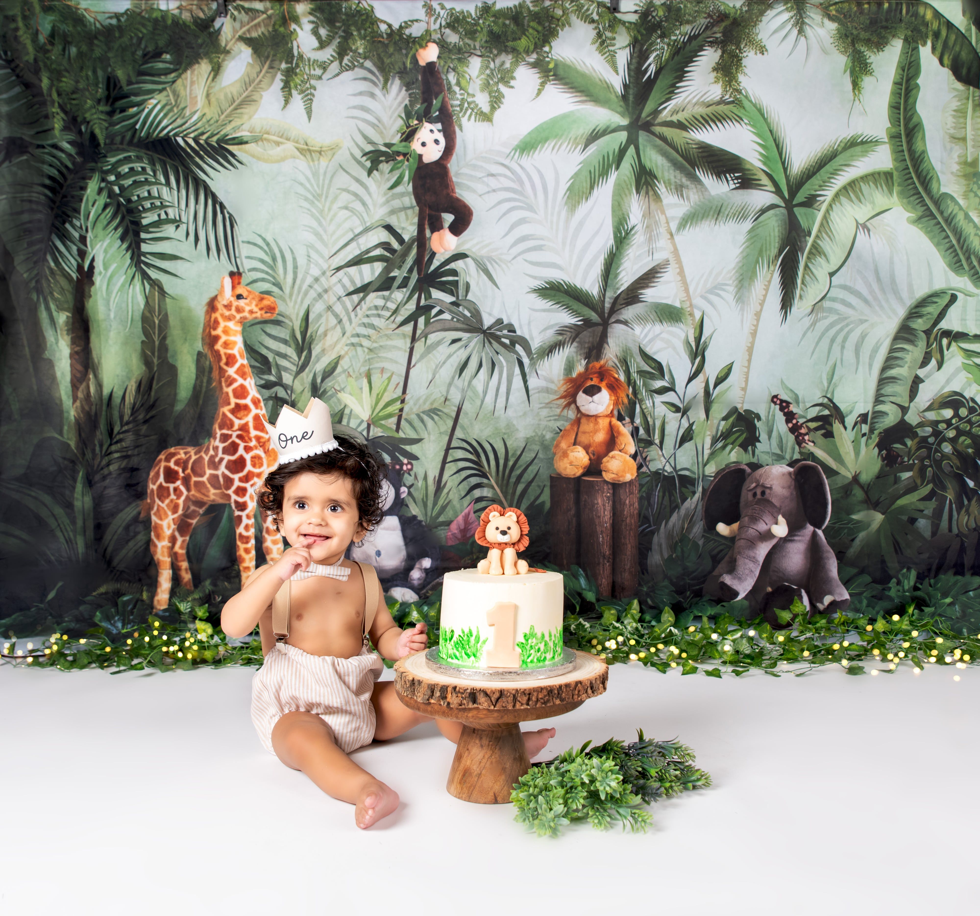 Toddler boy in tan romper sits beside jungle-themed first-birthday cake on safari studio set