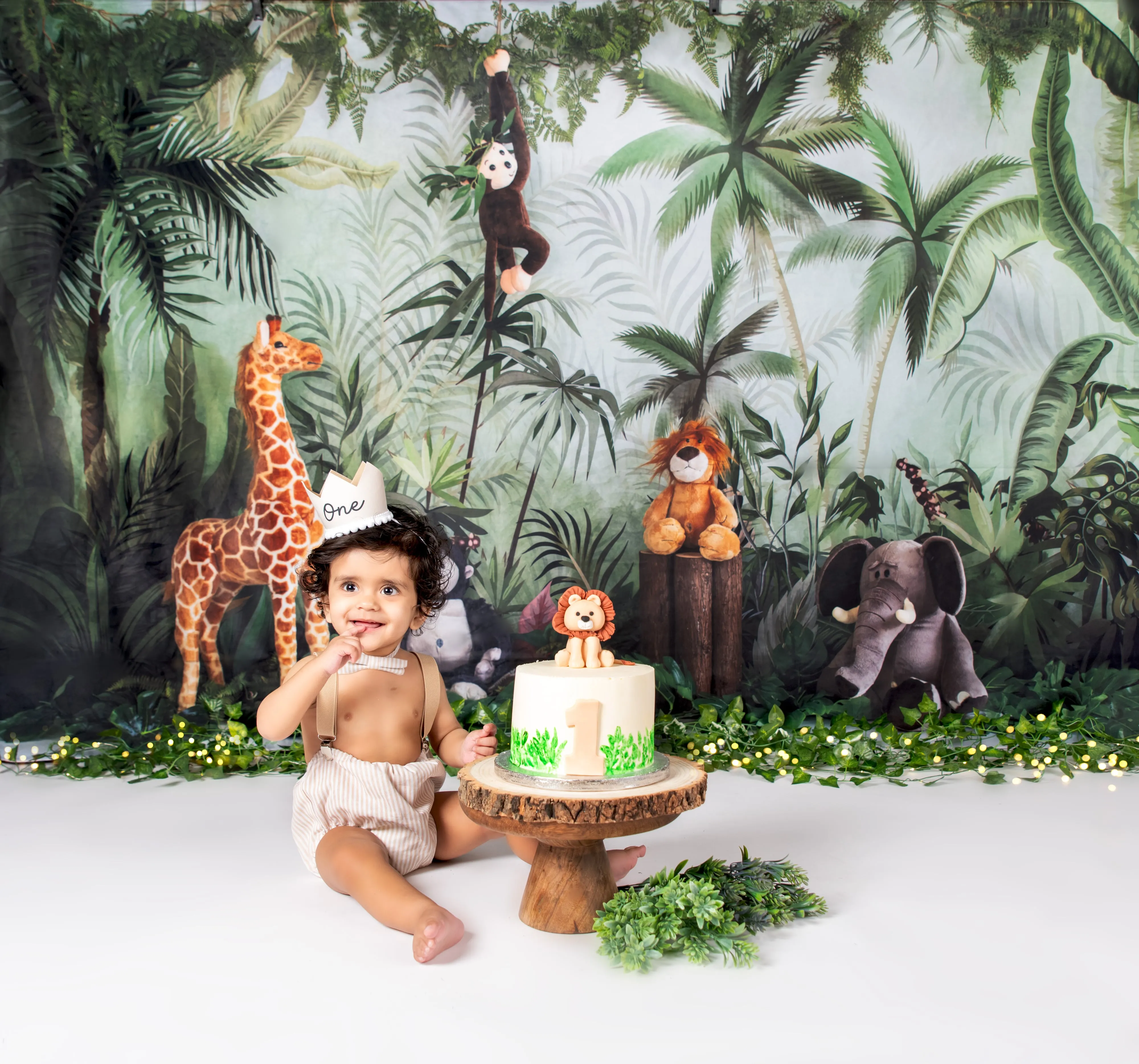 Toddler boy in tan romper sits beside jungle-themed first-birthday cake on safari studio set