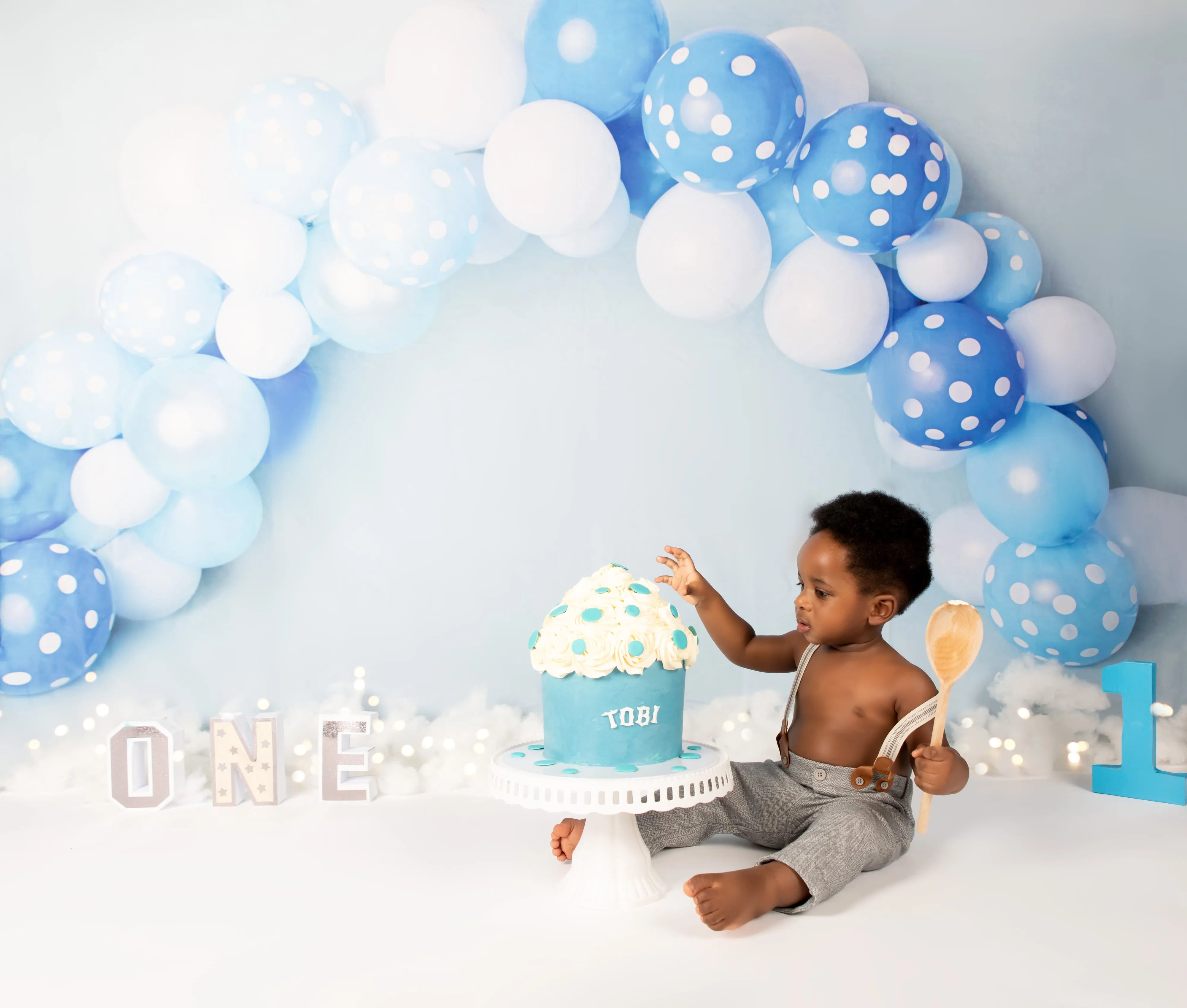 Toddler boy in grey shorts reaches for blue ombre TOBI cake under blue polka-dot balloon arch
