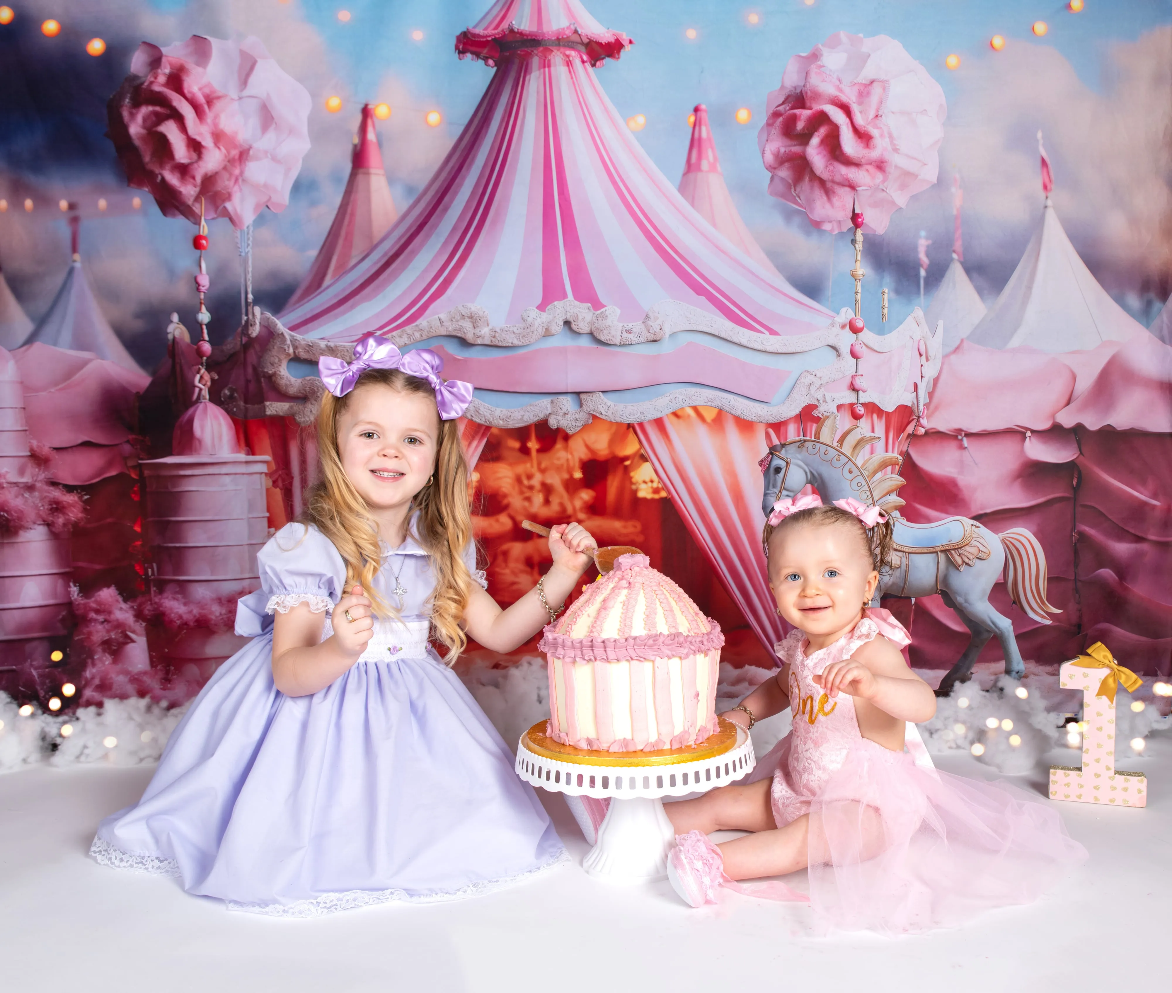Two sisters in lilac and pink share carousel-themed cake on circus-tent studio backdrop