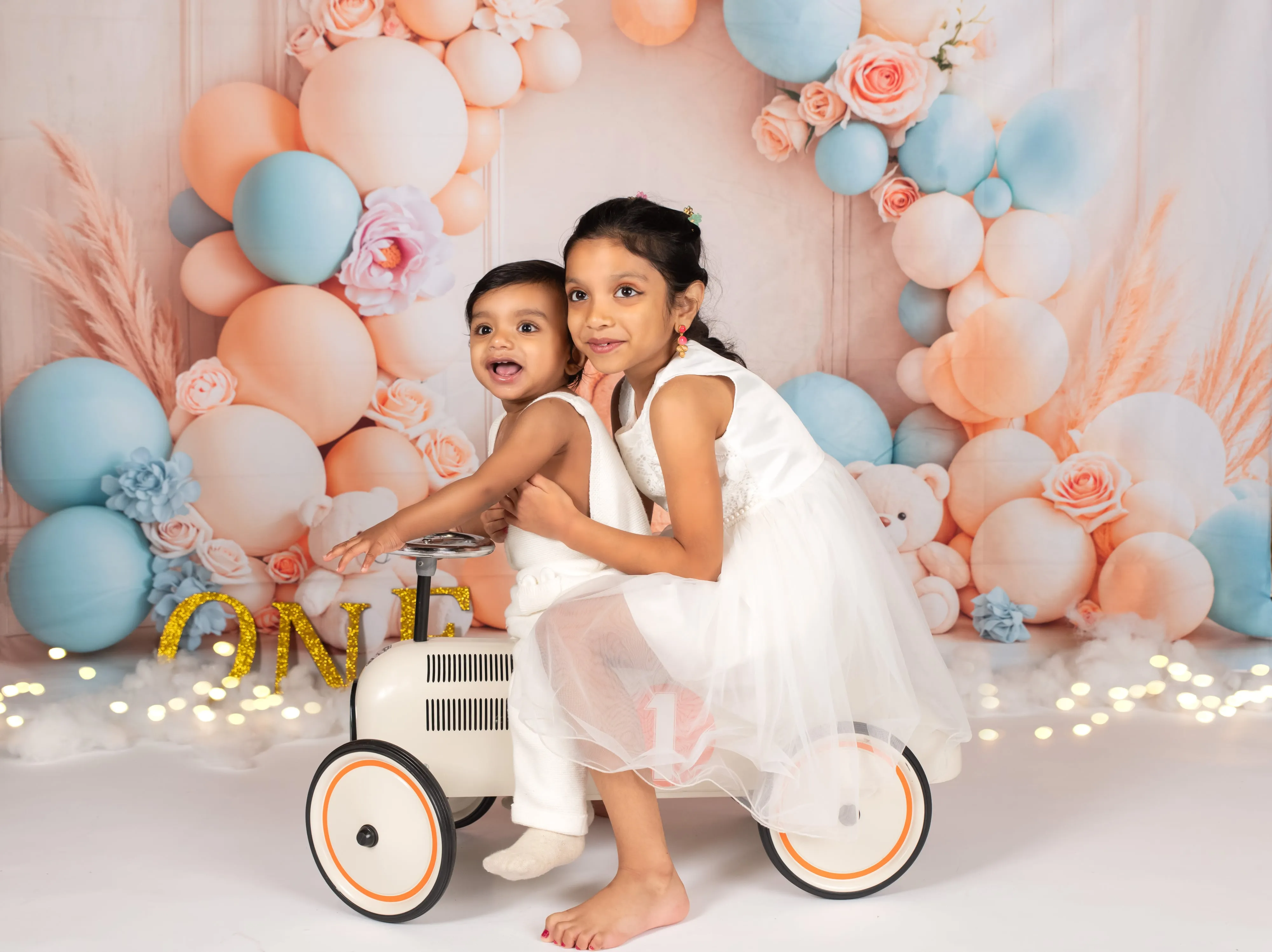 Two sisters share white toy car on peach-and-blue pastel balloon arch with florals
