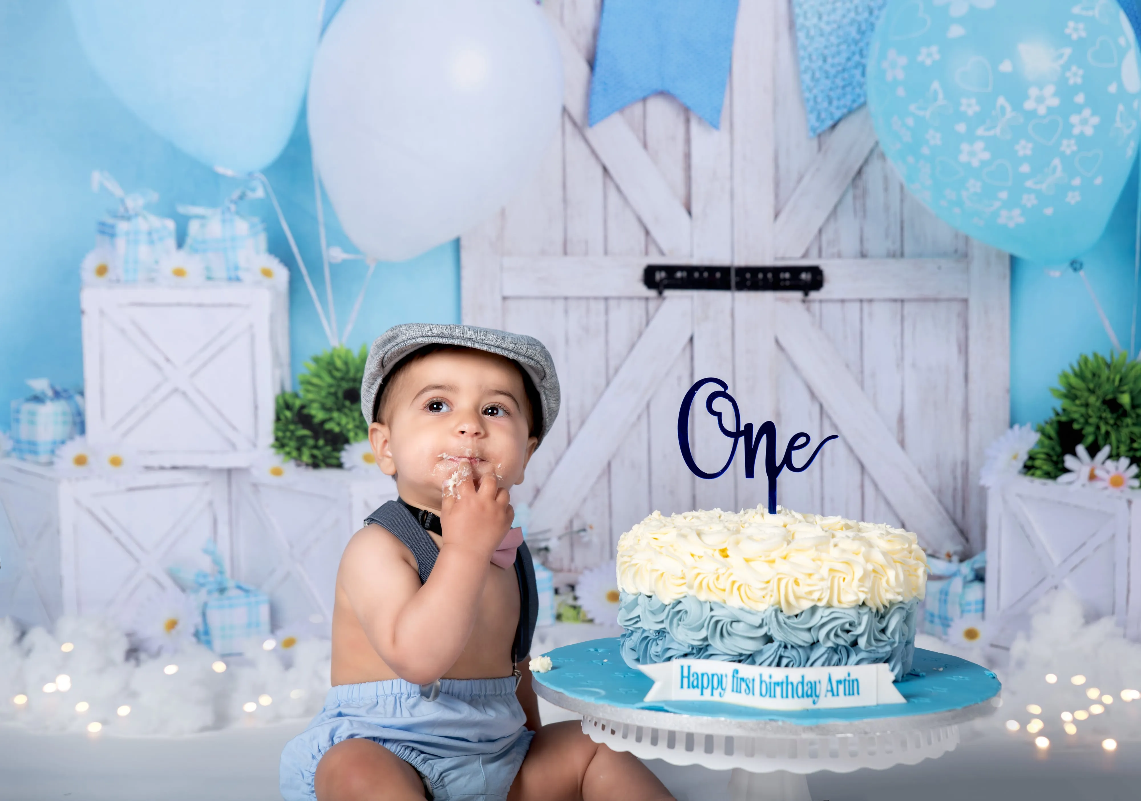 Toddler boy in flat cap and dungarees beside ombre Arin cake on rustic blue barn-door set