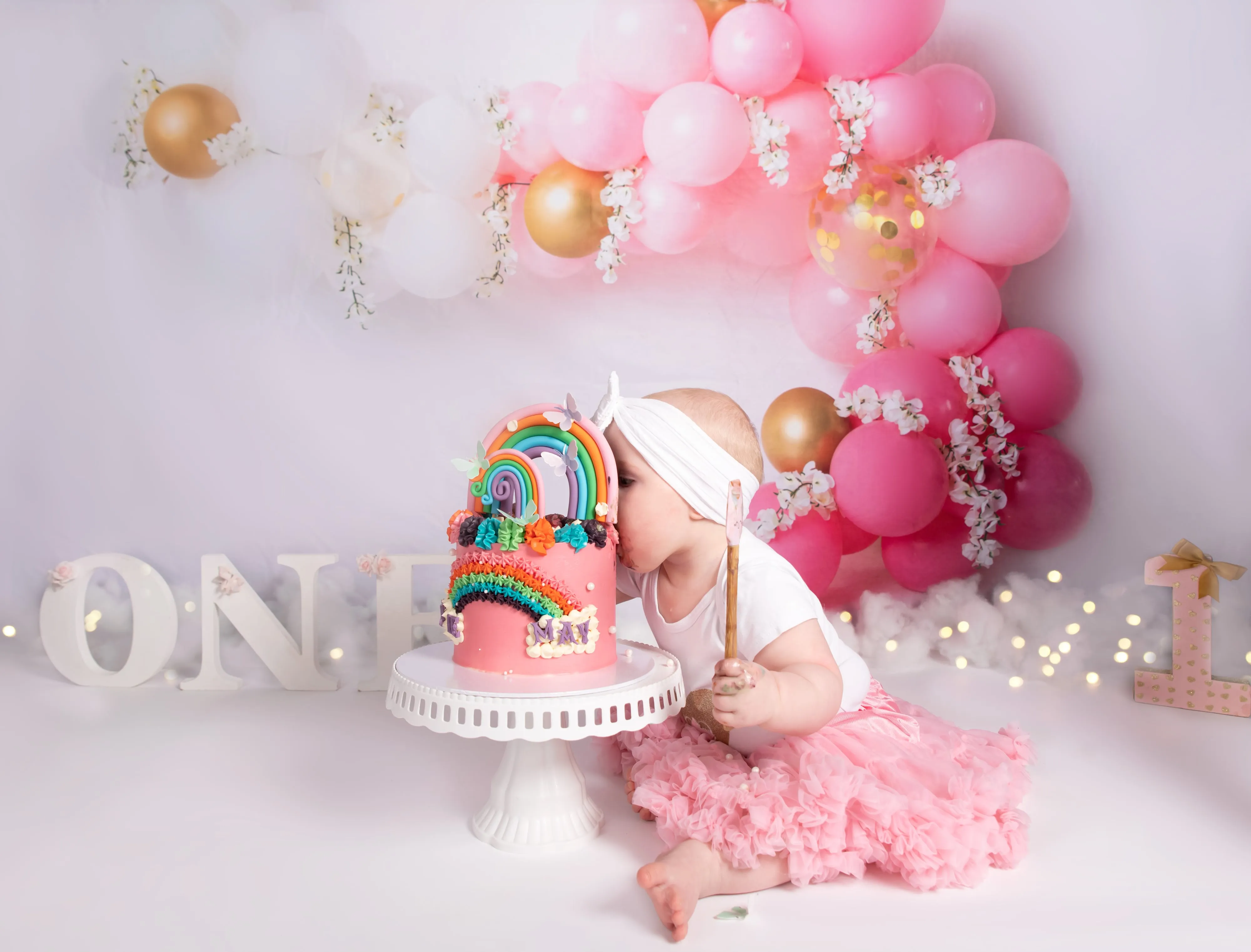 Toddler in pink tutu kisses unicorn rainbow cake under pink and gold balloon arch