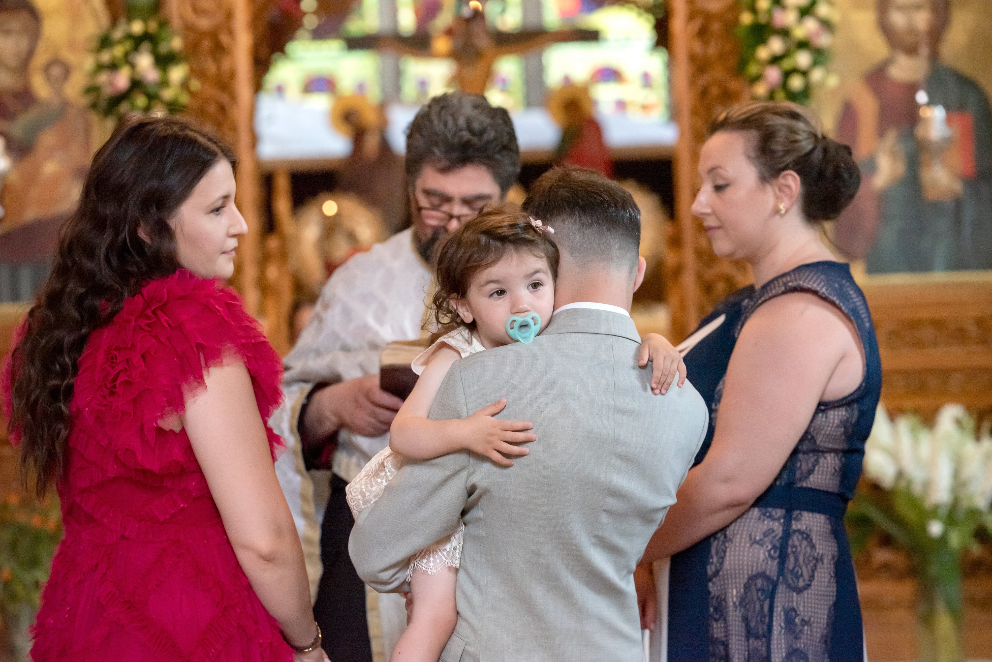 Toddler held close during an Orthodox christening ceremony, family gathered before the gilded iconostasis