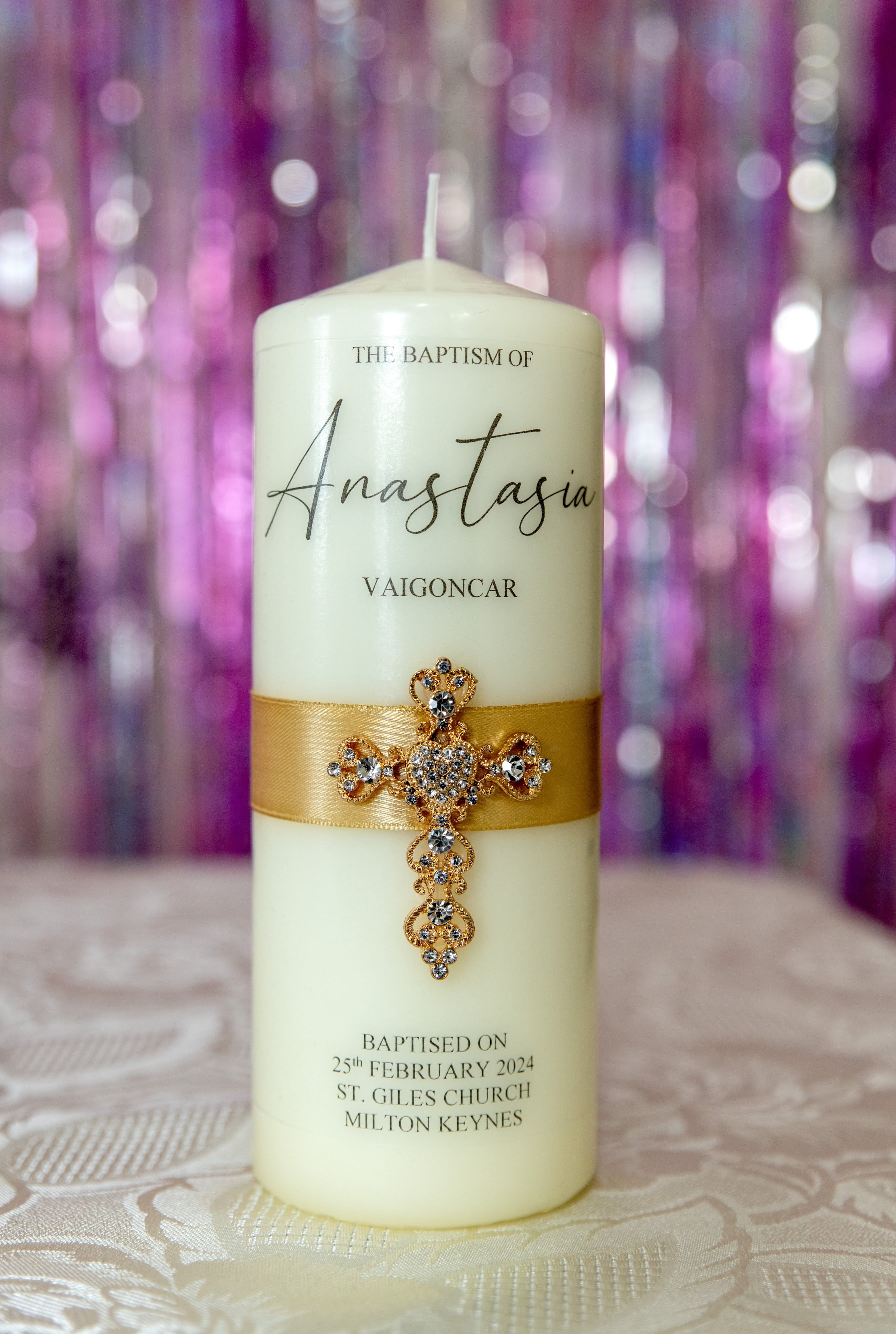 Personalised baptism candle tied with a jewelled gold cross, set against a soft pink shimmer backdrop