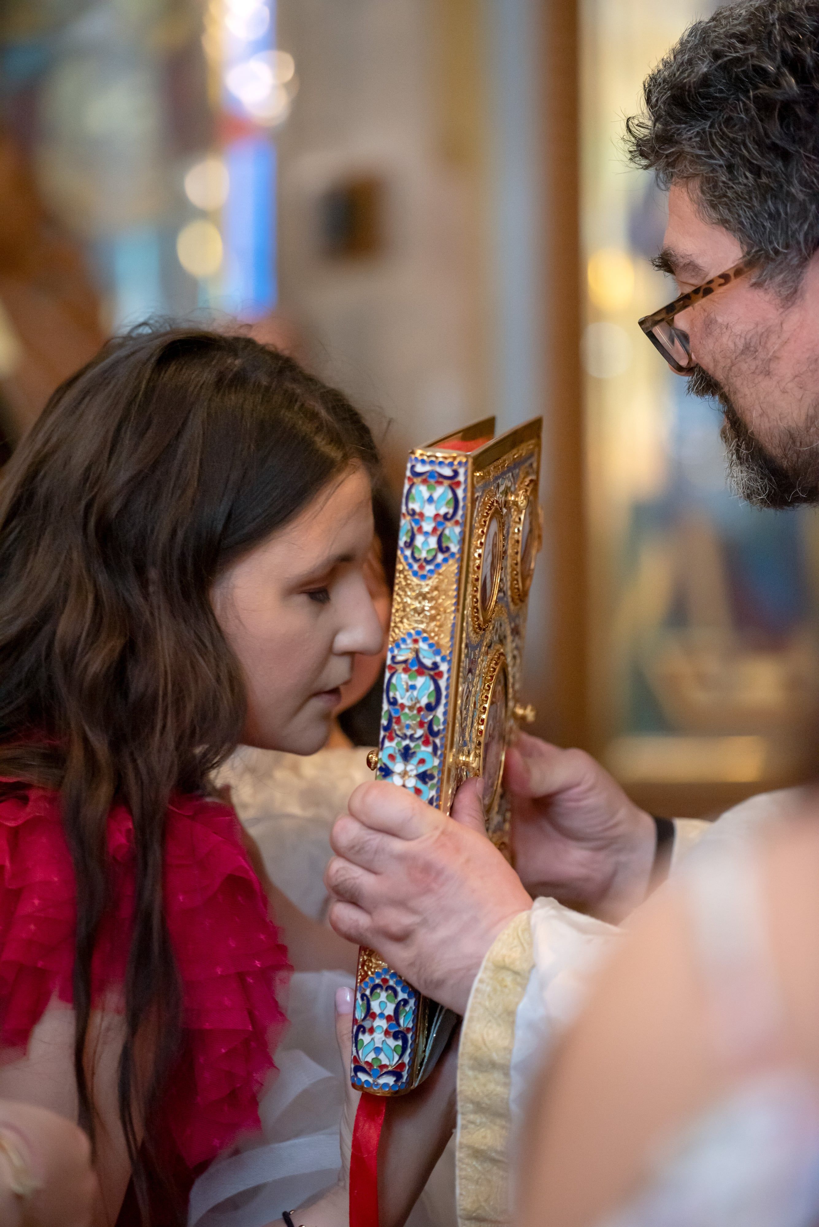 Young girl in a red dress kisses the ornate gilded gospel book held by the priest at an Orthodox christening