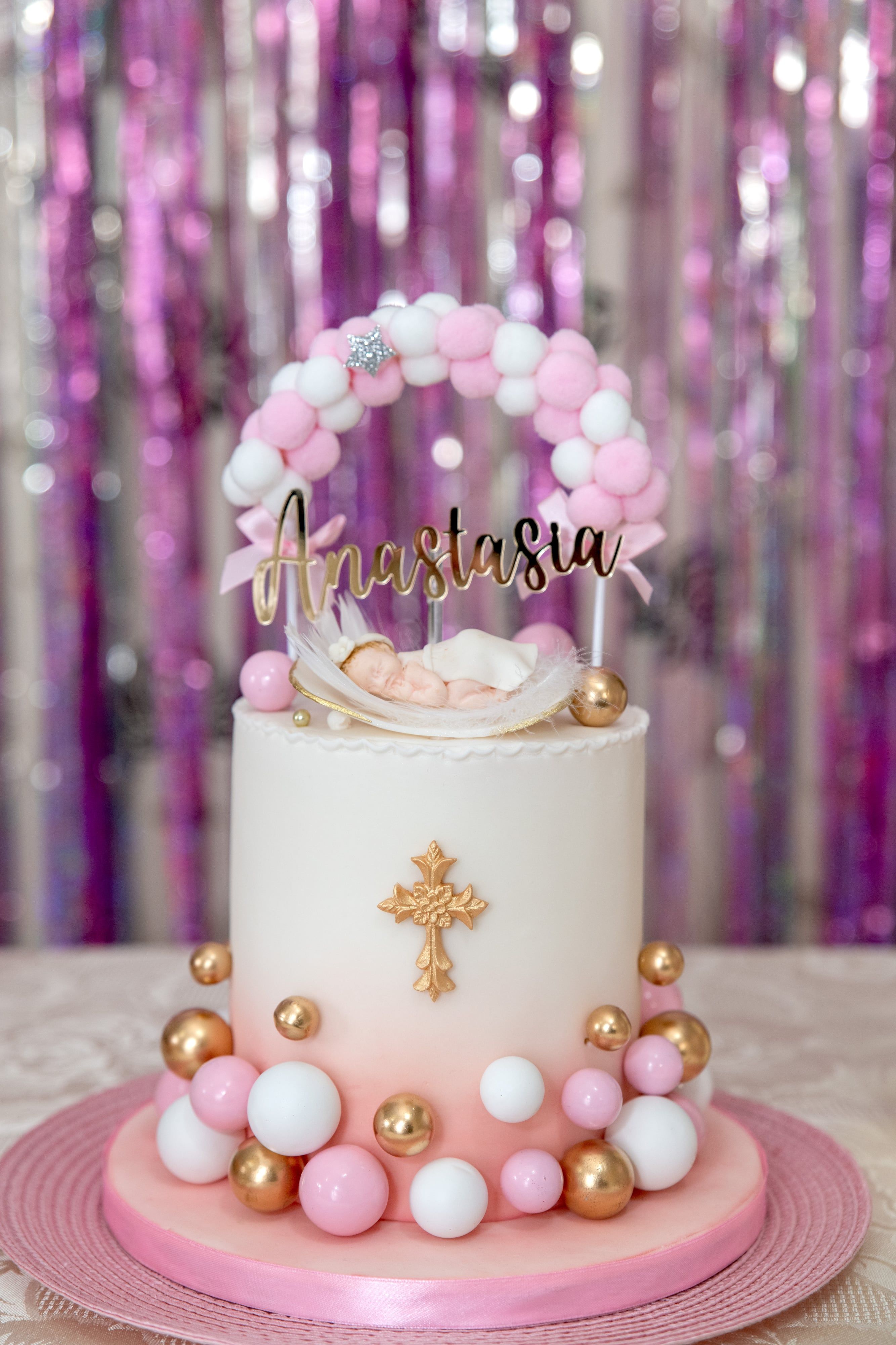 Pink-and-gold christening cake topped with a balloon arch and gilded cross beneath a shimmer backdrop