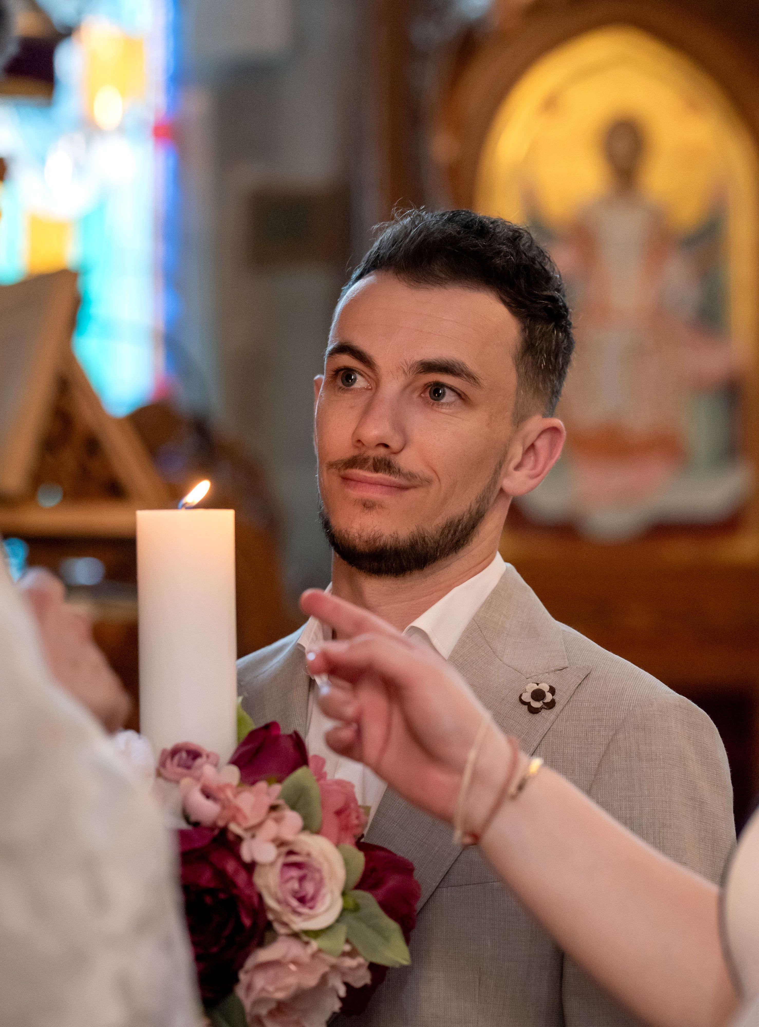 A man holds the lit baptism candle and roses during an Orthodox christening, icons glowing behind him