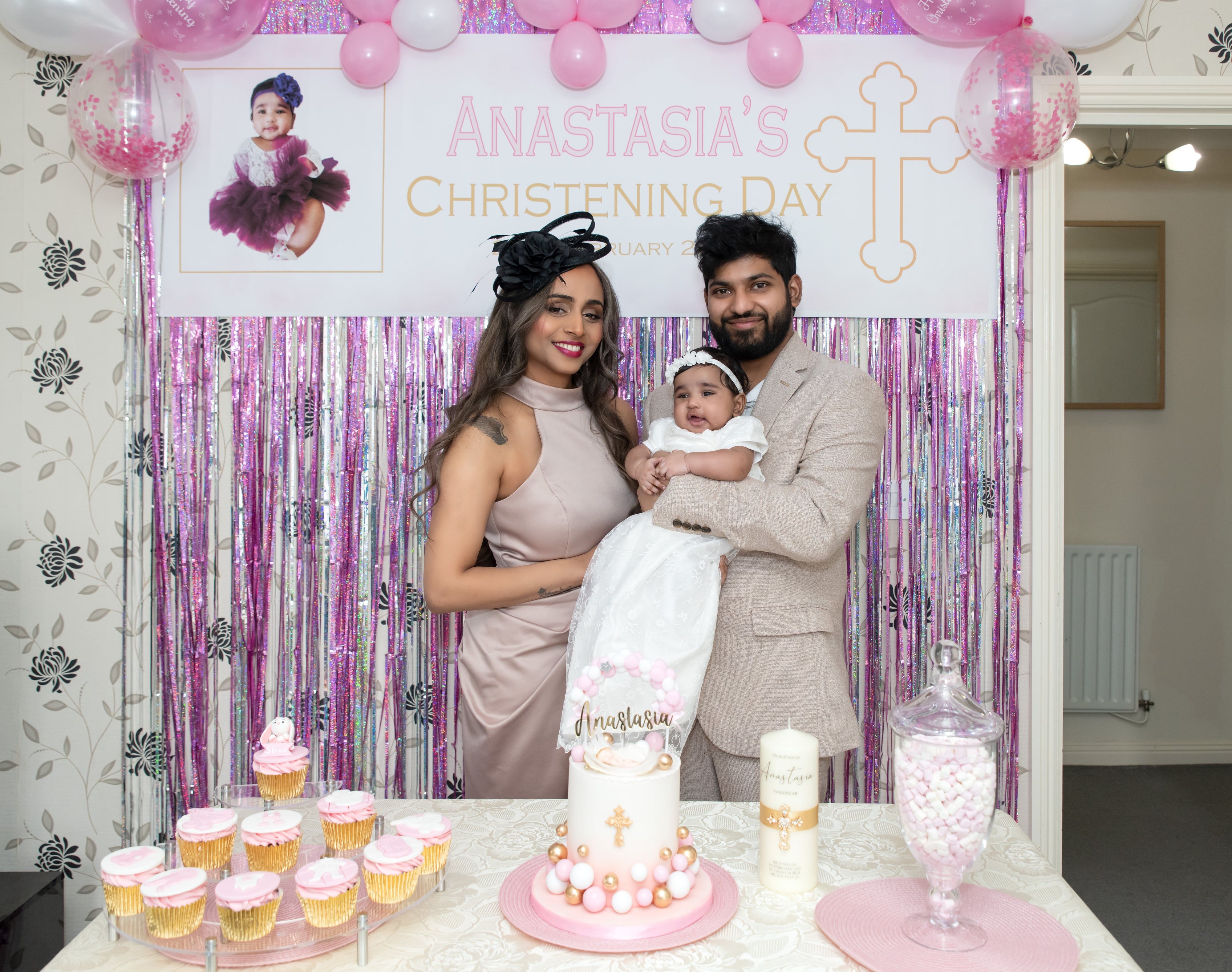 Parents hold their baby in a white gown before a christening-day backdrop, dessert table dressed in pink