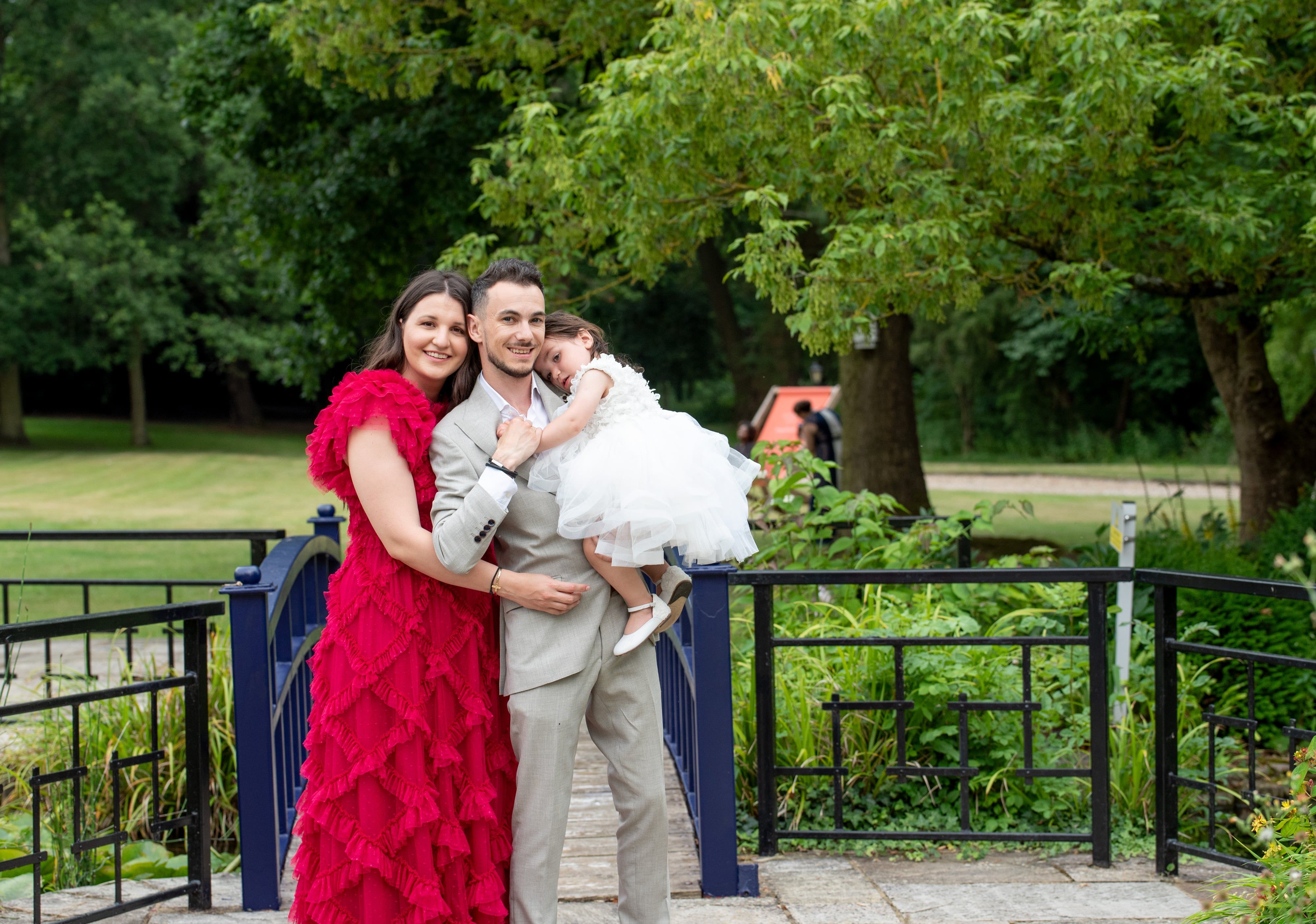 Family of three on an ornate blue garden bridge, their toddler in a white tulle christening dress