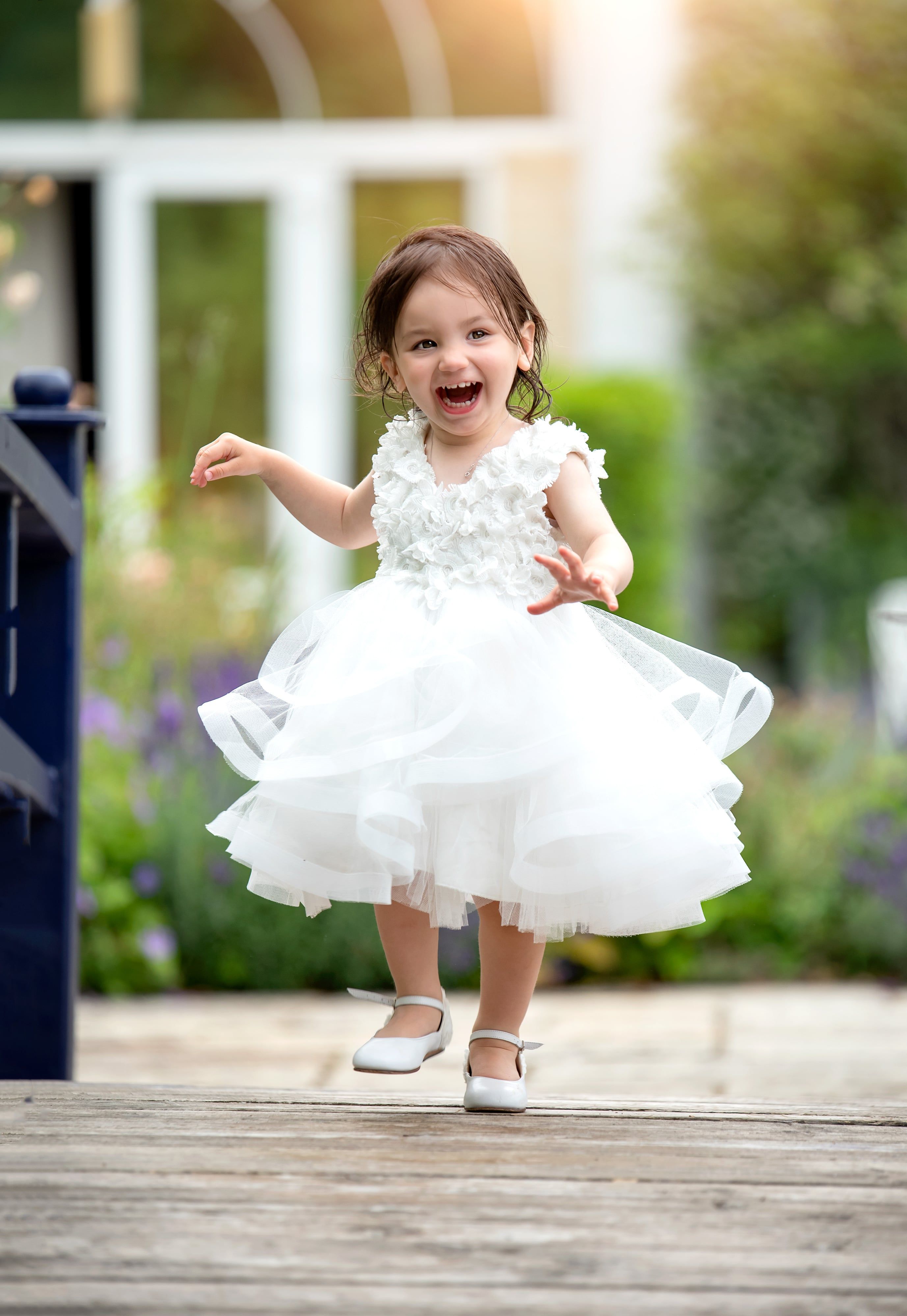Toddler in a white tulle dress laughing mid-run along garden decking after her christening