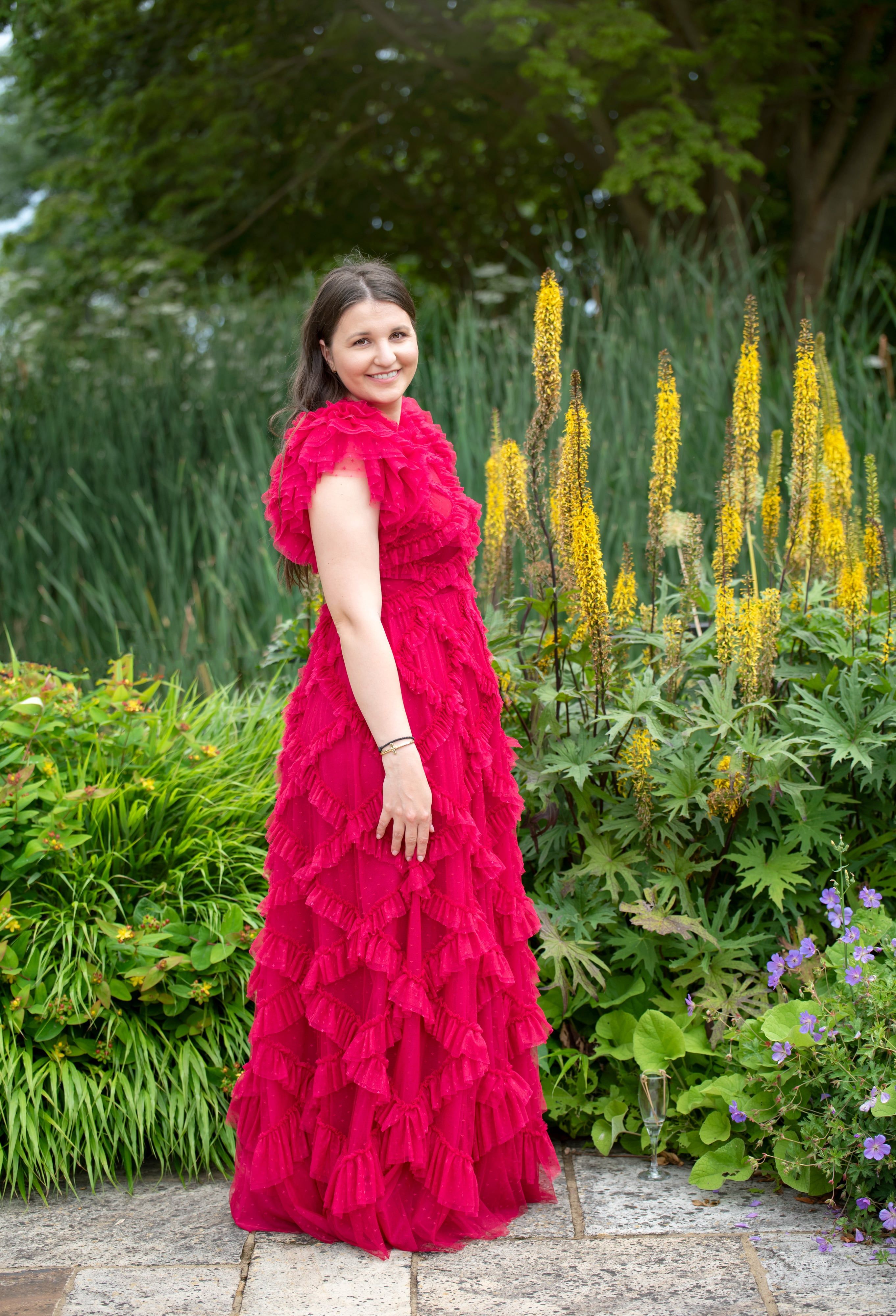 Mother in a flowing red ruffled gown turns to smile among tall yellow garden spires