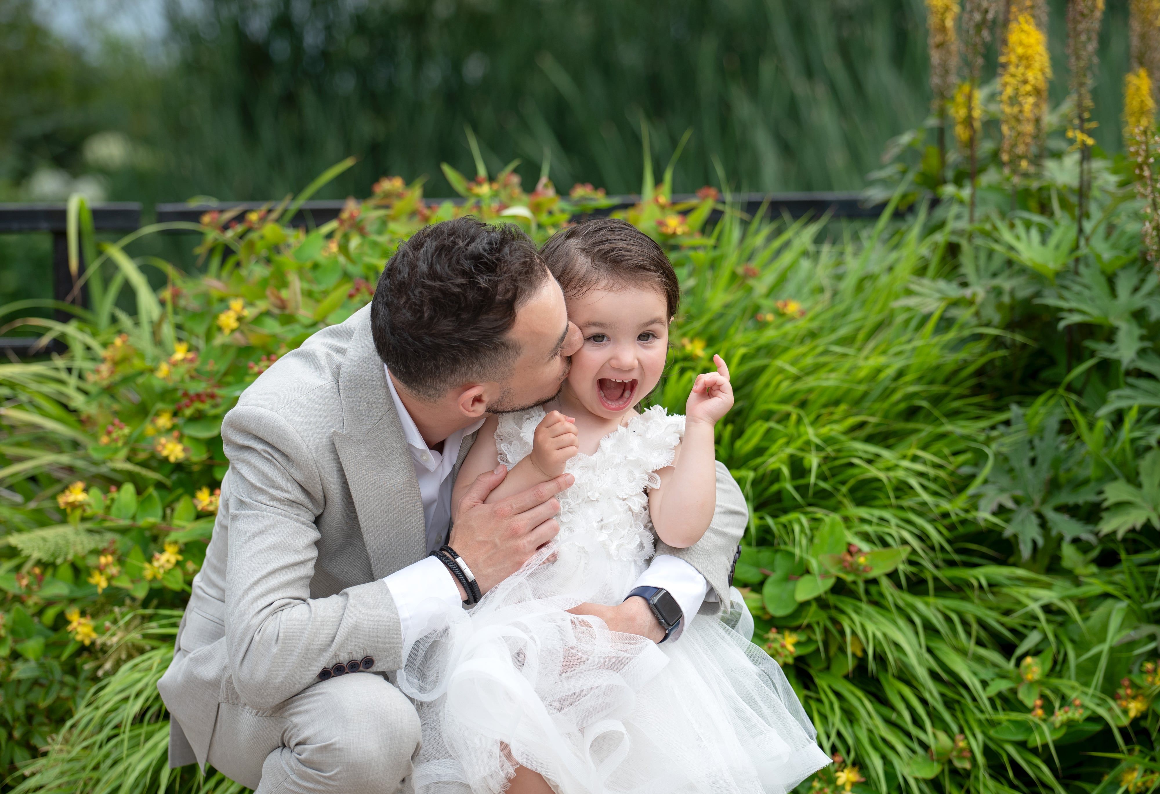 Father kisses his delighted toddler's cheek among garden greenery after her christening