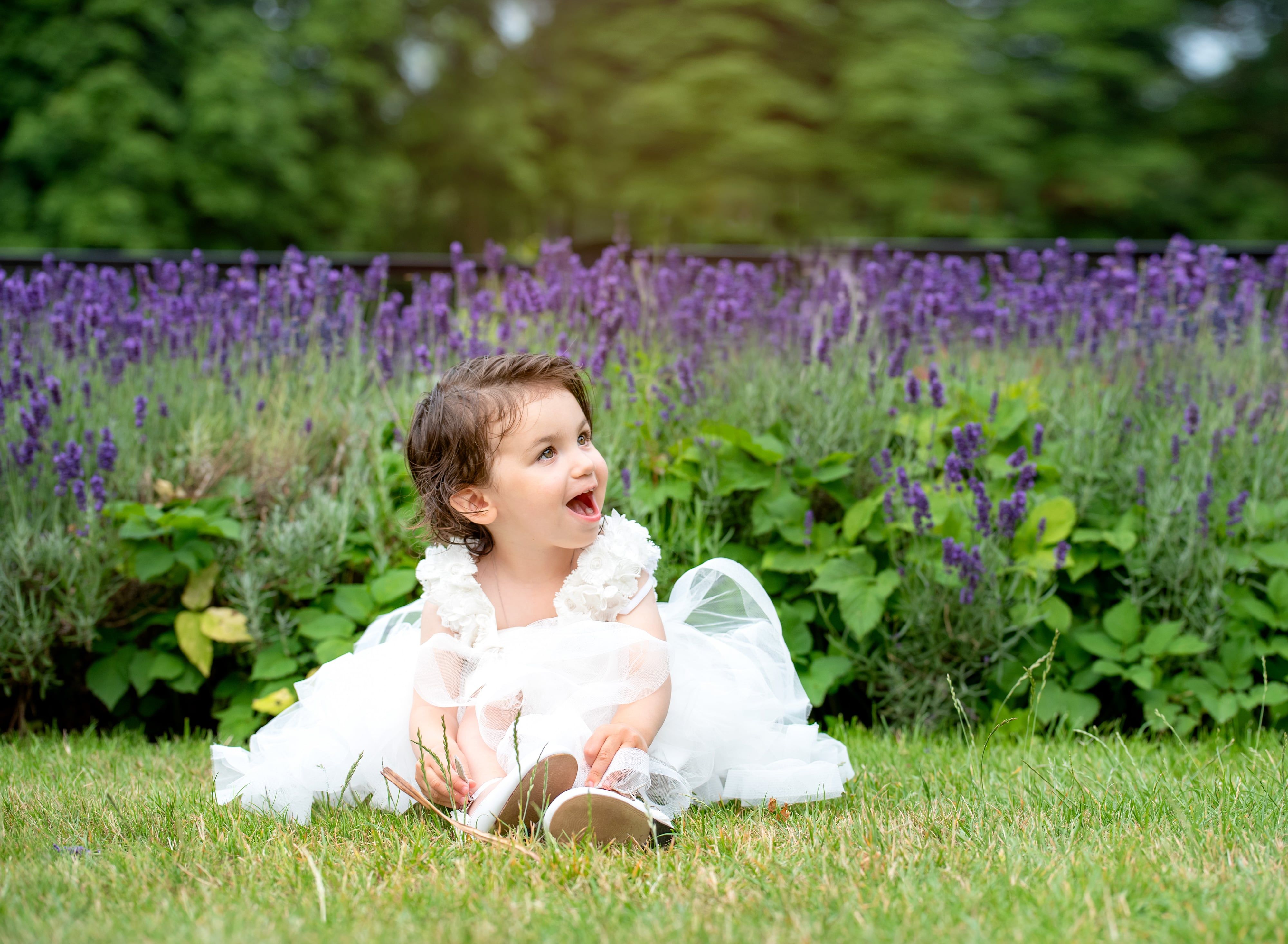Toddler in a white christening dress sits on the grass beside lavender, caught mid-delight