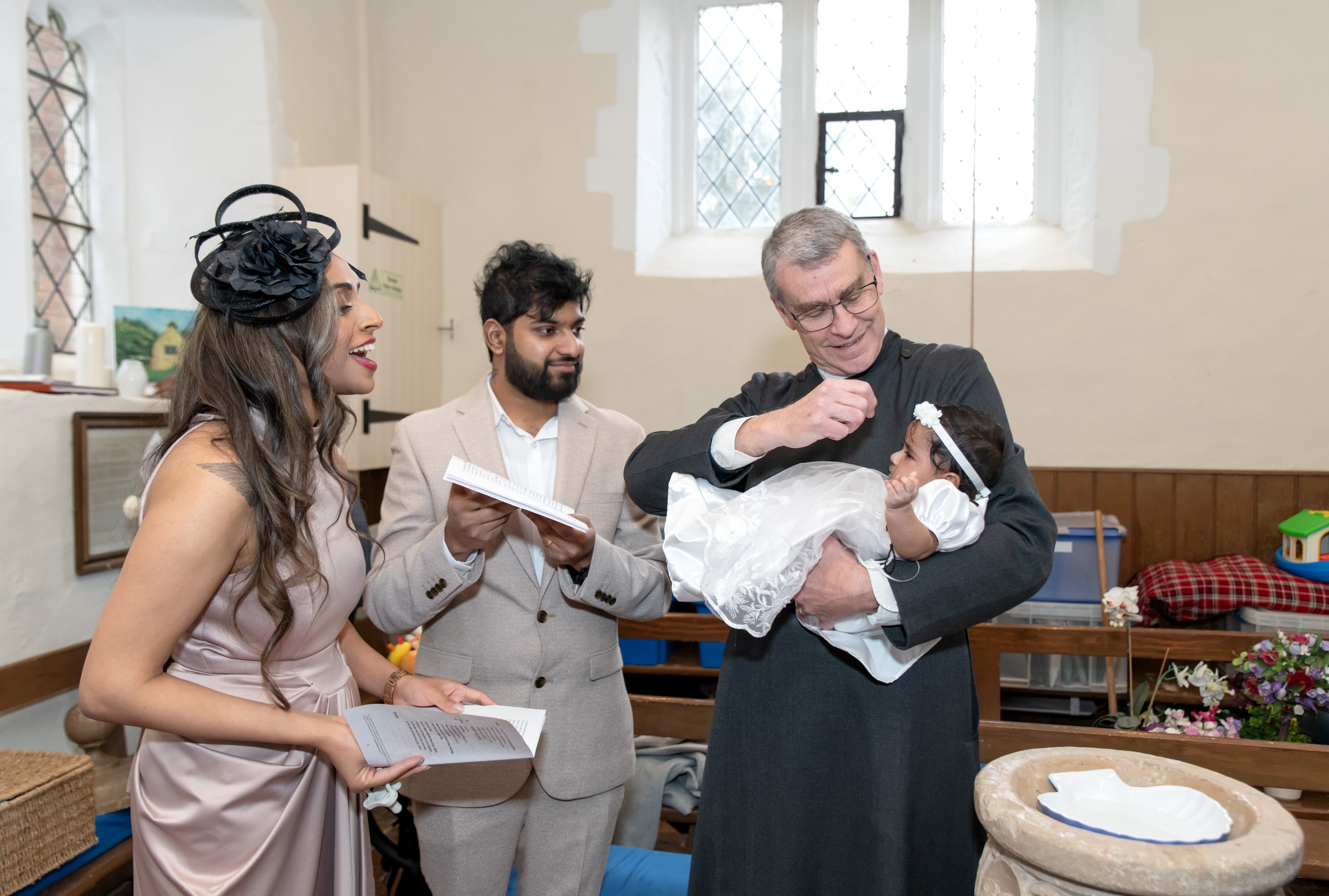 Vicar marks the baby with the sign of the cross as her mother laughs with joy beside the font