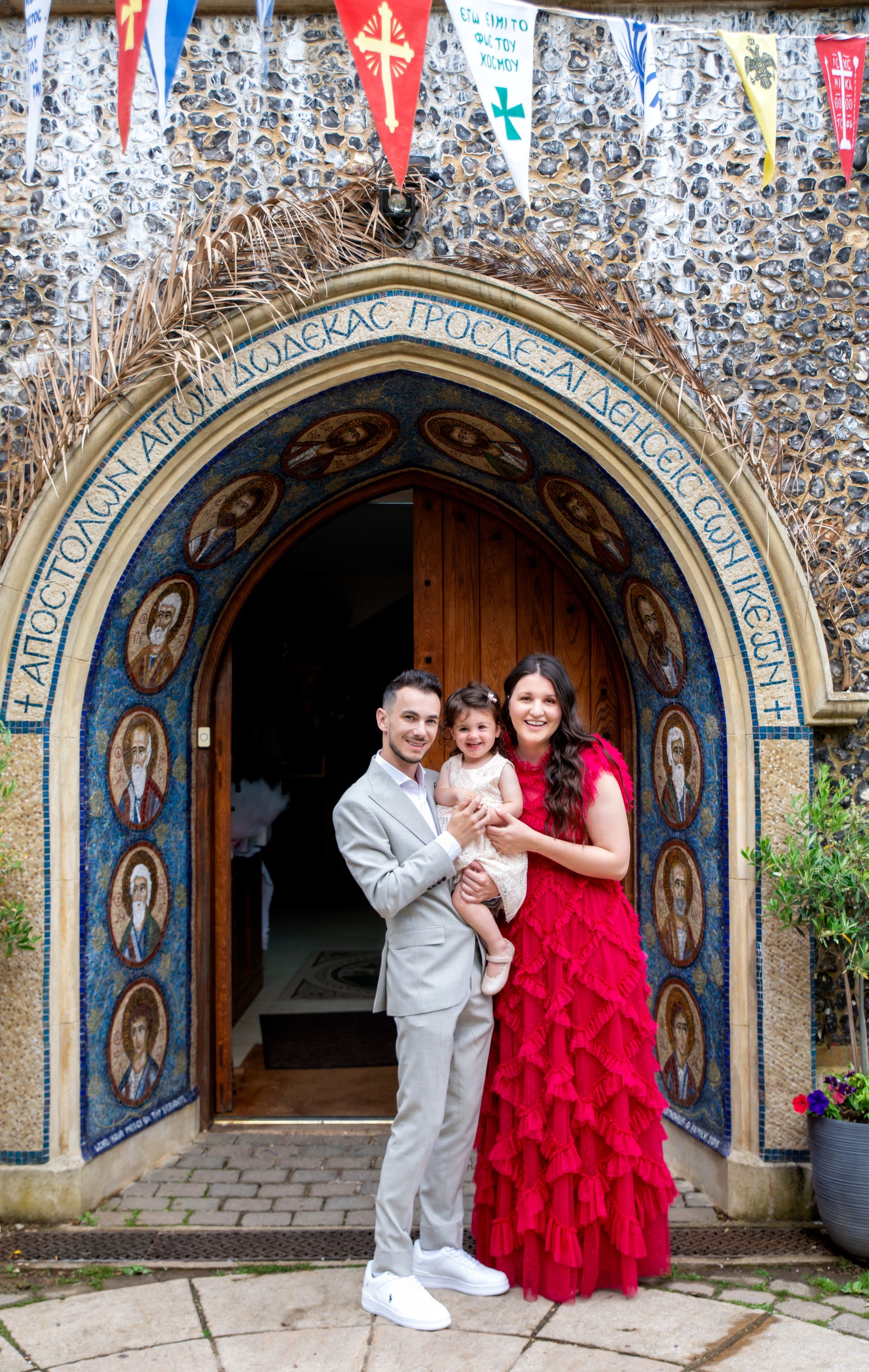 Family of three at the mosaic-tiled arched doorway of an Orthodox church, bunting strung above