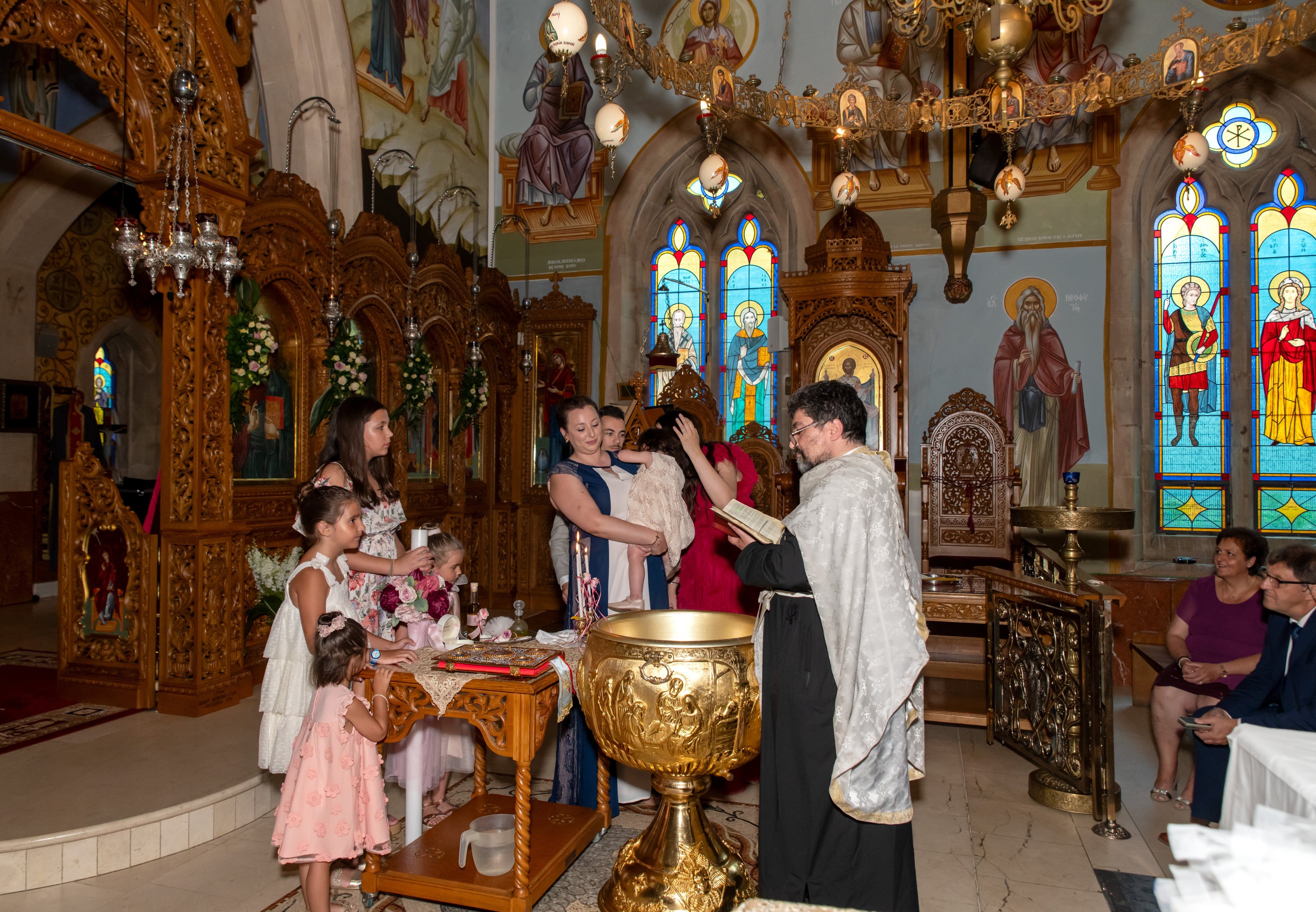 Priest performs the baptism at an ornate gold font as two small flower girls look on