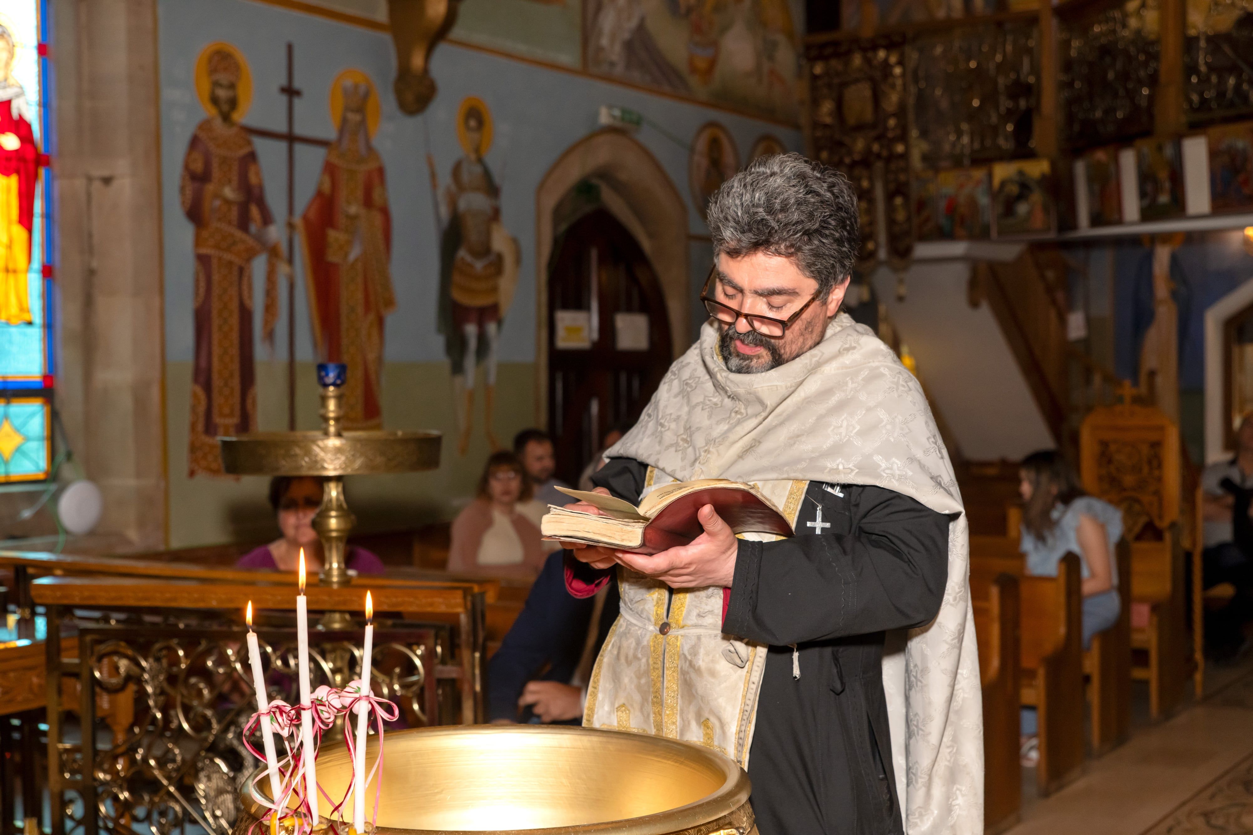 Priest reads from the gospel beside the gold font and twin baptism candles during an Orthodox christening
