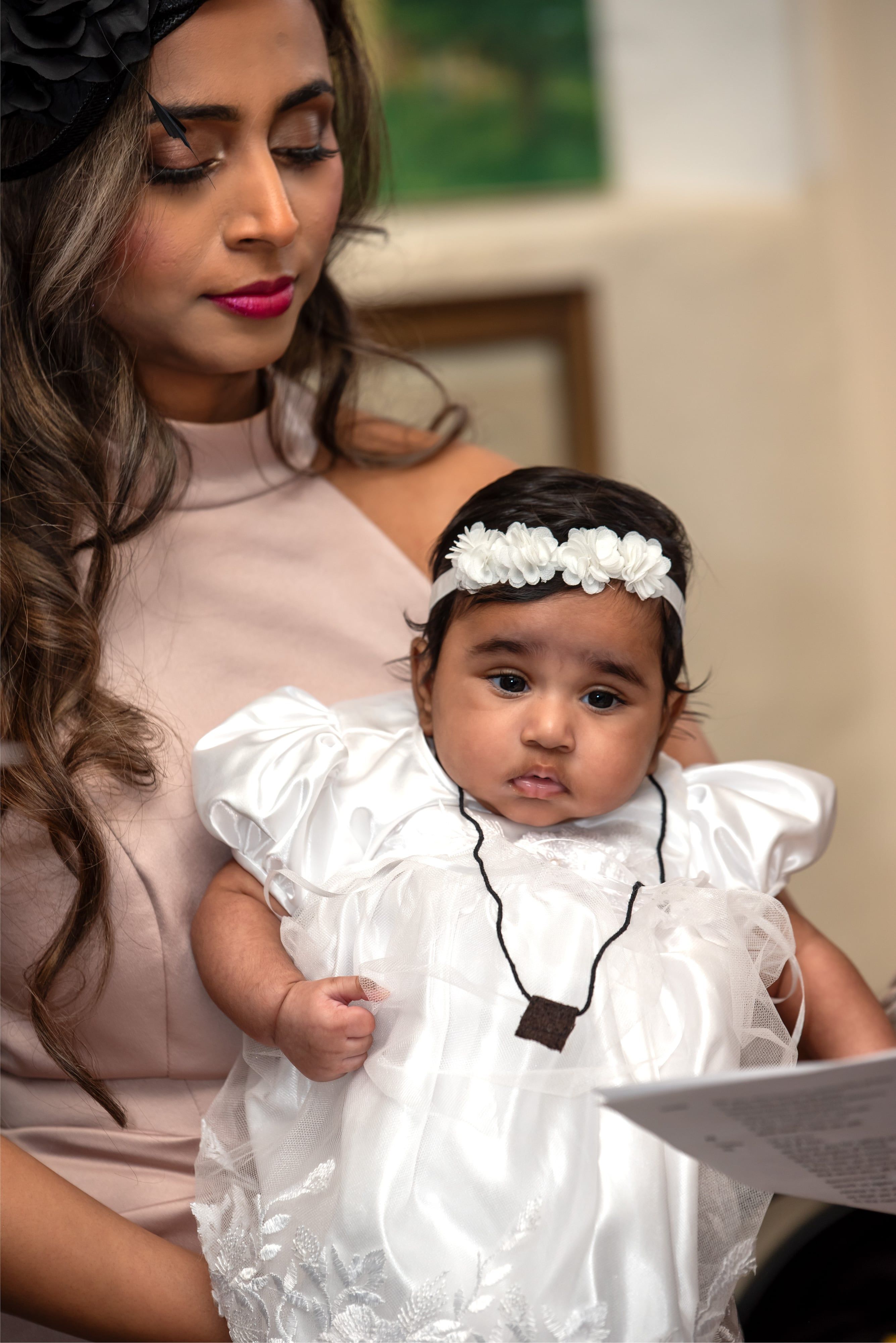 Mother in a fascinator holds her baby in a white gown and floral headband during the christening